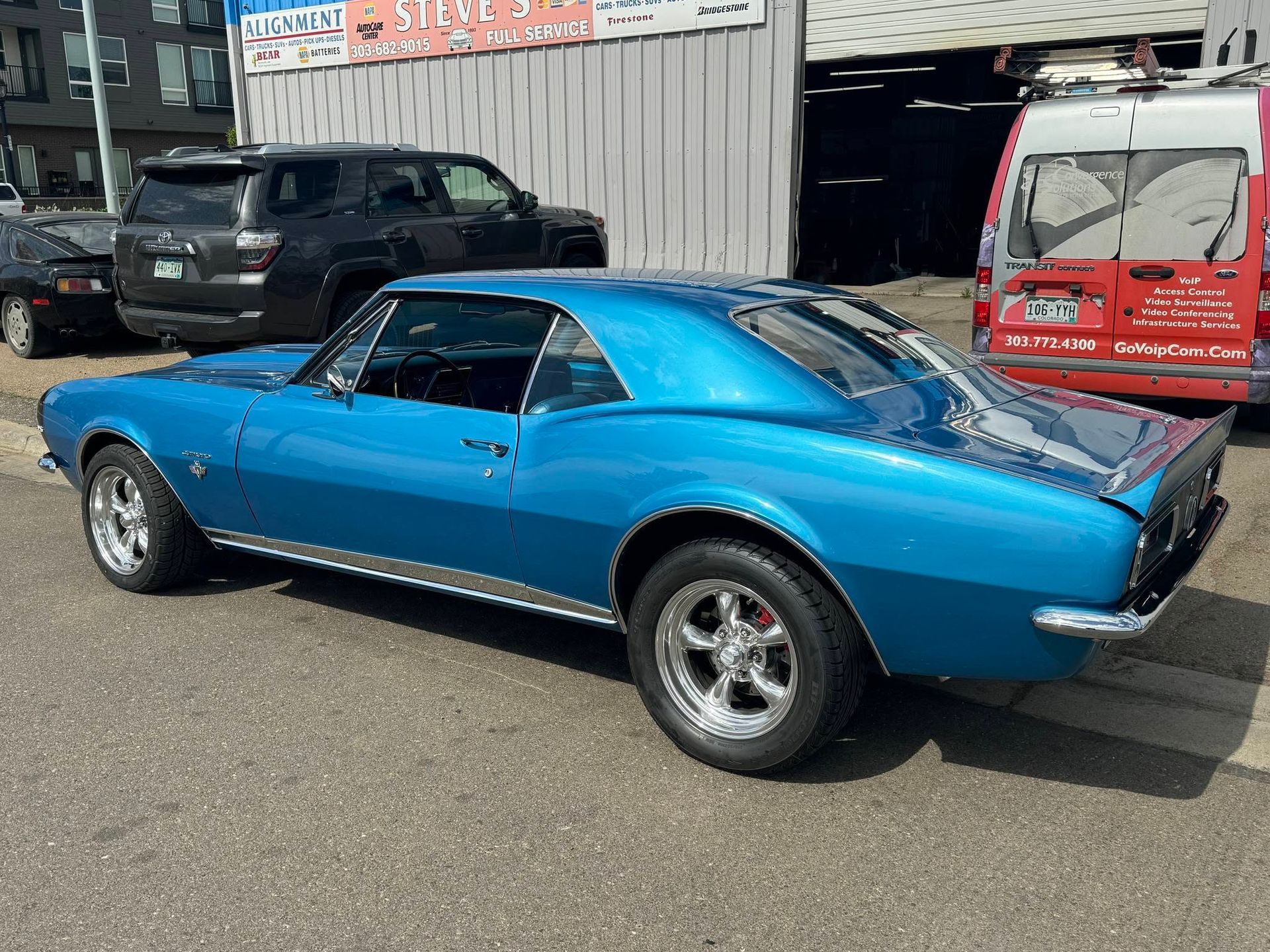 Blue classic muscle car parked outside an auto shop with a red van in the background