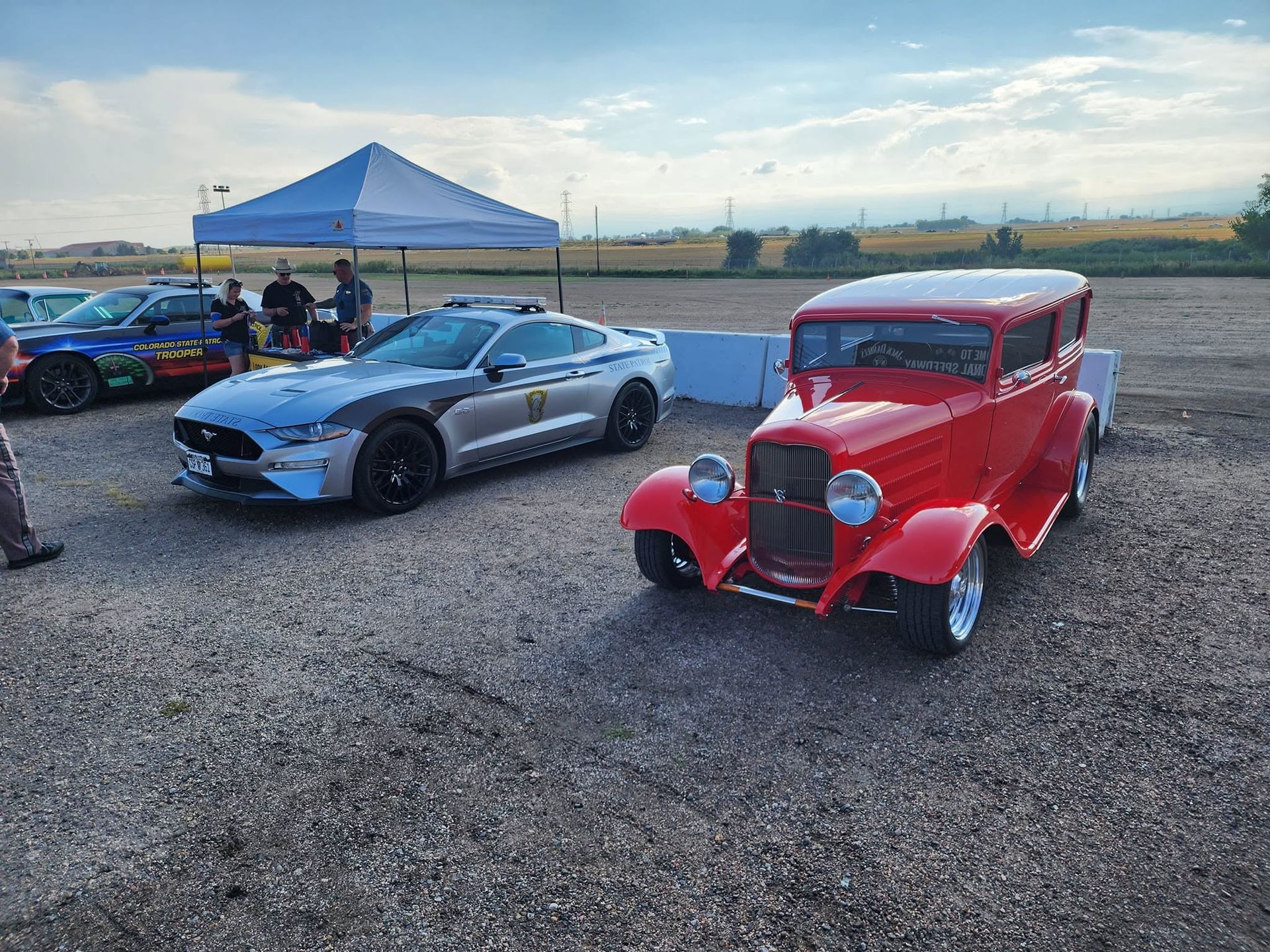 Classic red hot rod and silver sports car parked on gravel near a blue canopy at sunset.