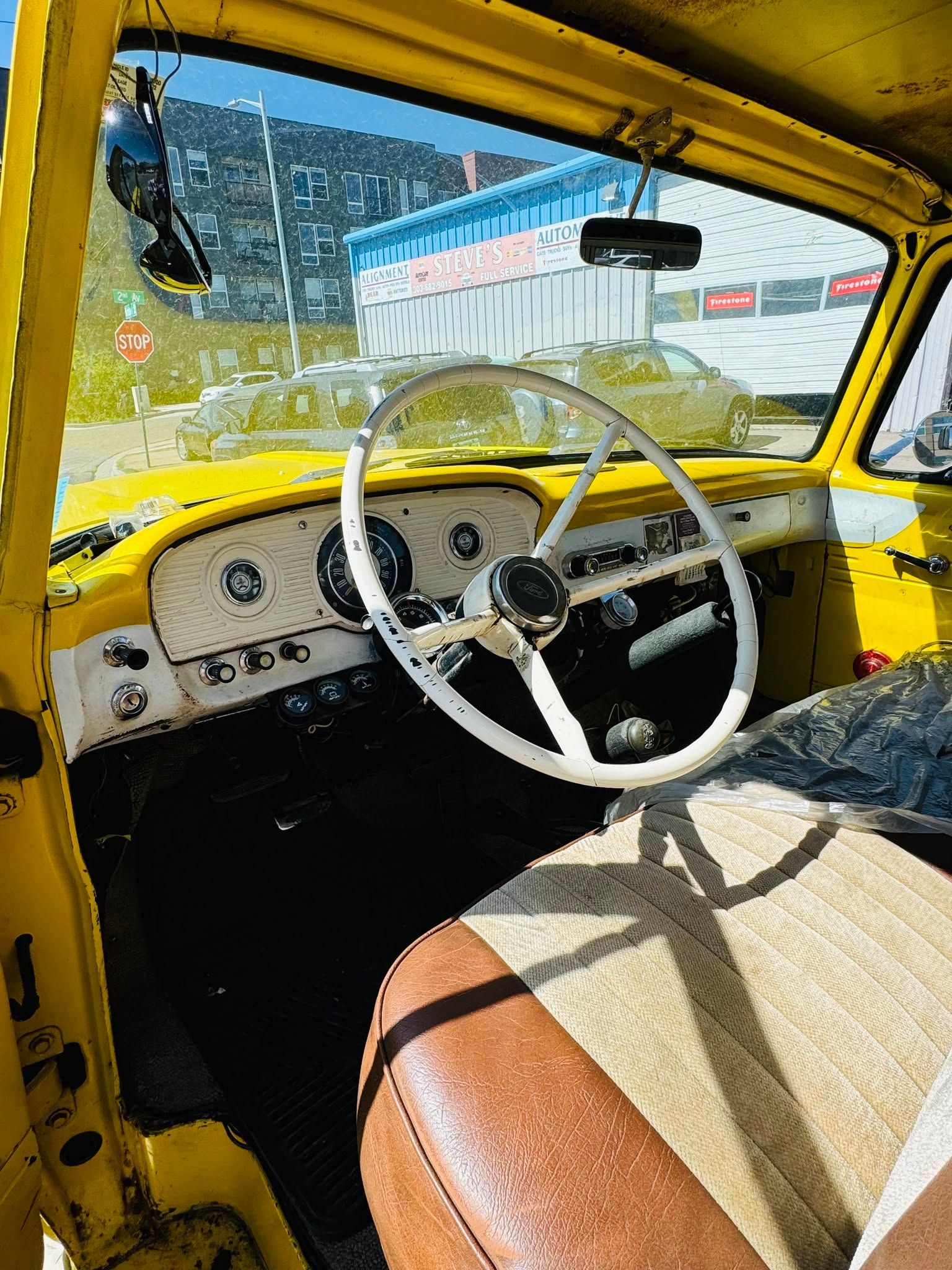 Interior of a vintage yellow car with a white steering wheel and plaid front seat, viewed from the driver’s side