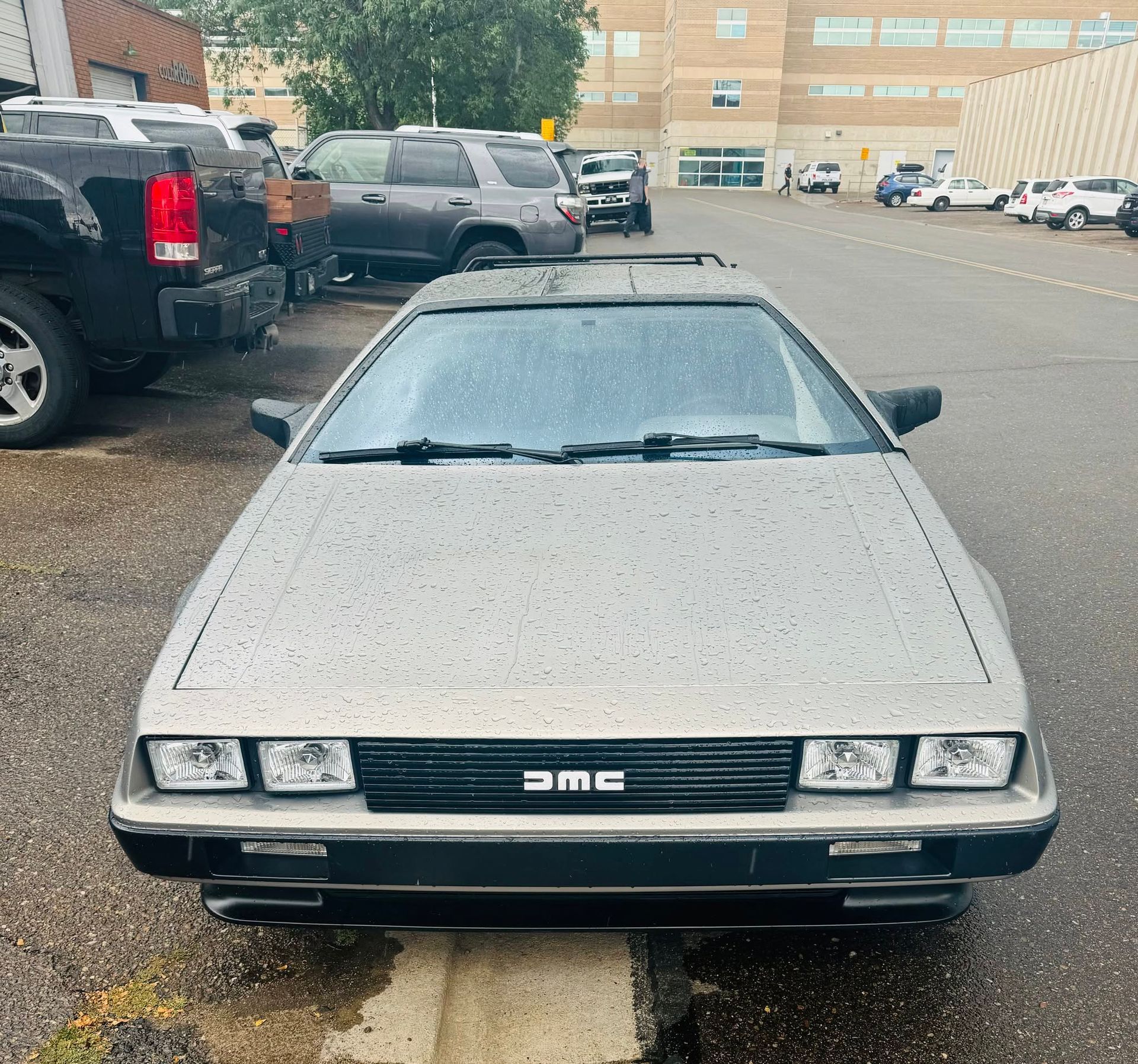 Silver DeLorean DMC-12 parked in a lot, front view on a wet street.