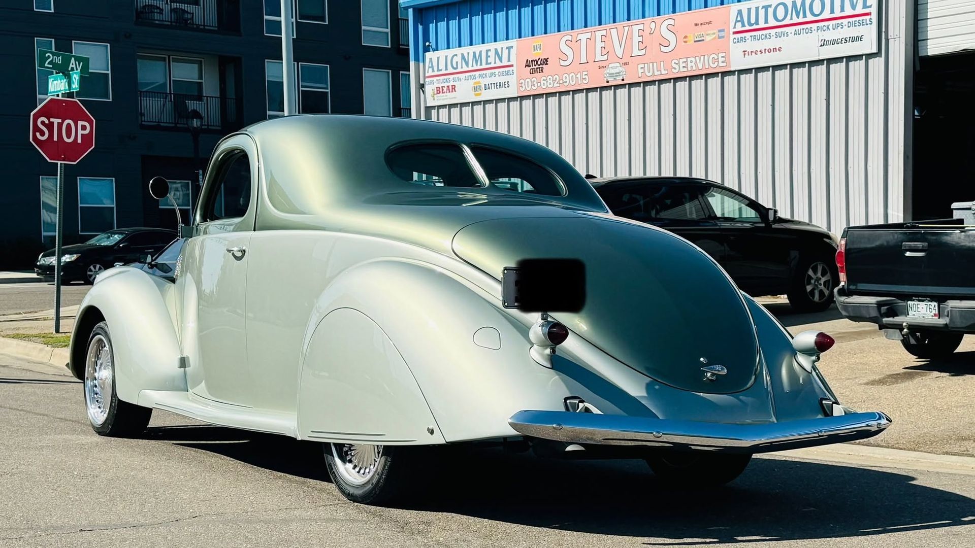 Vintage silver coupe parked on a street beside an auto shop sign and a stop sign