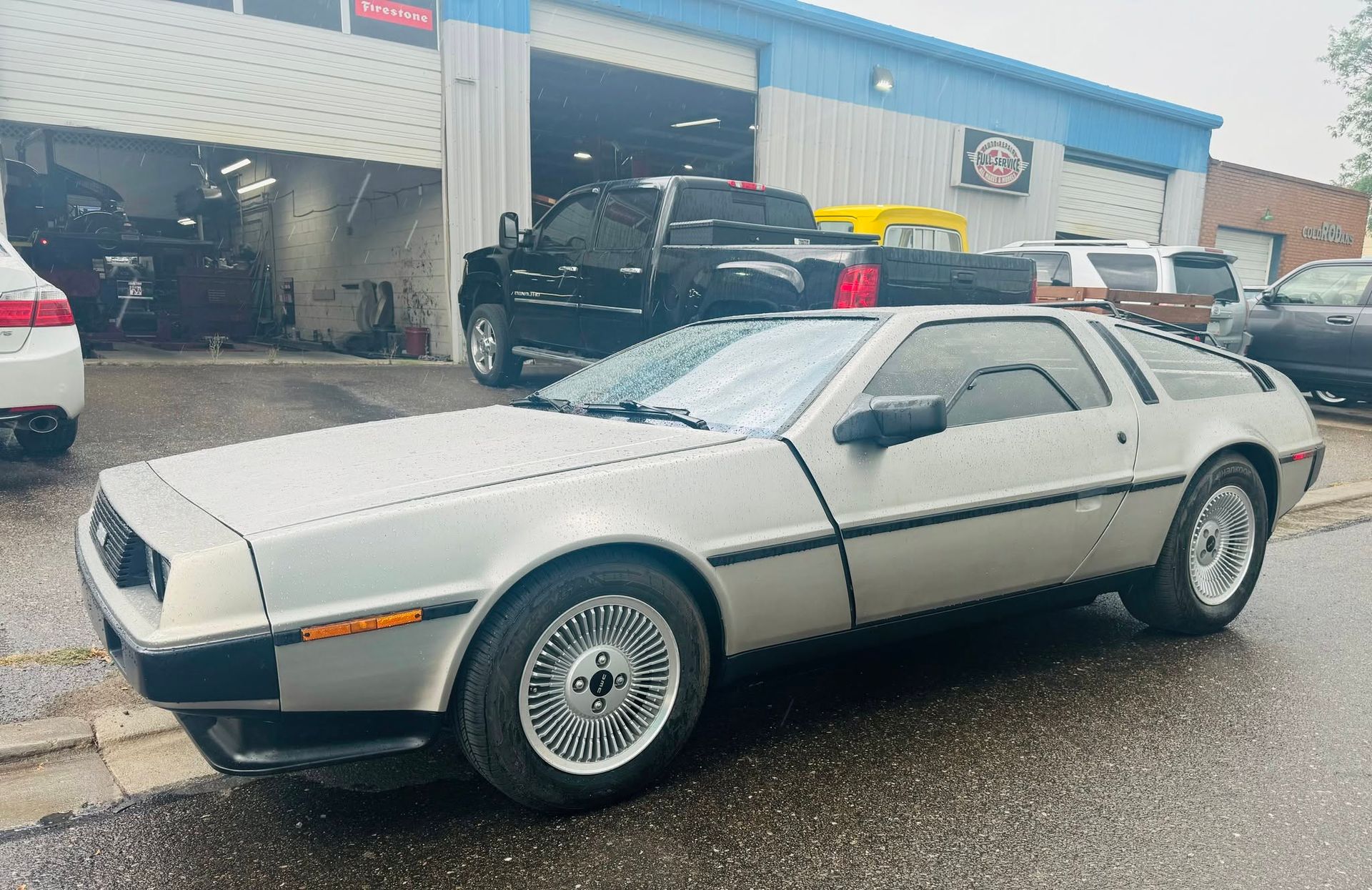Silver DeLorean sports car parked on a wet street outside a building under overcast skies