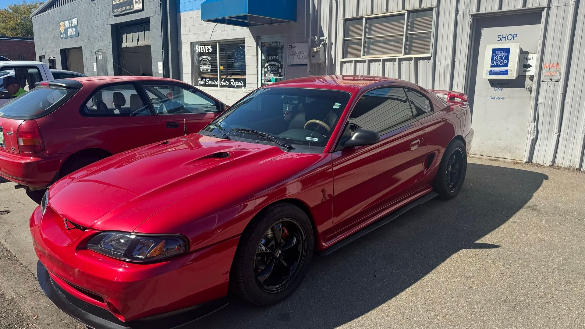 Red sports coupe parked outside a building on a sunny day, with another red car beside it.
