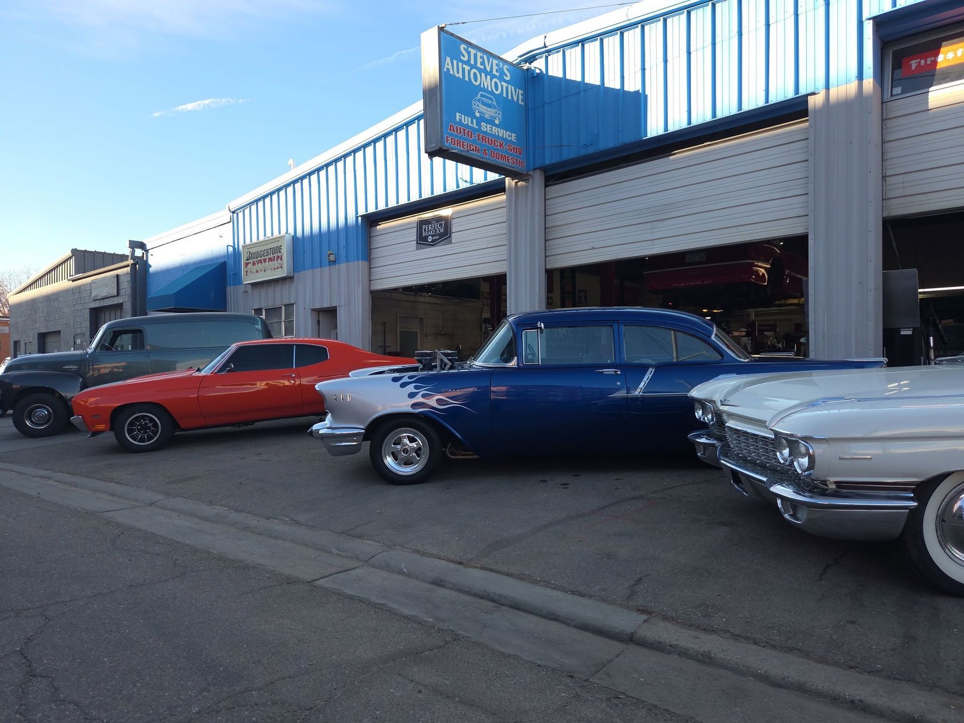 Classic cars parked outside a garage with open bays under a blue awning.
