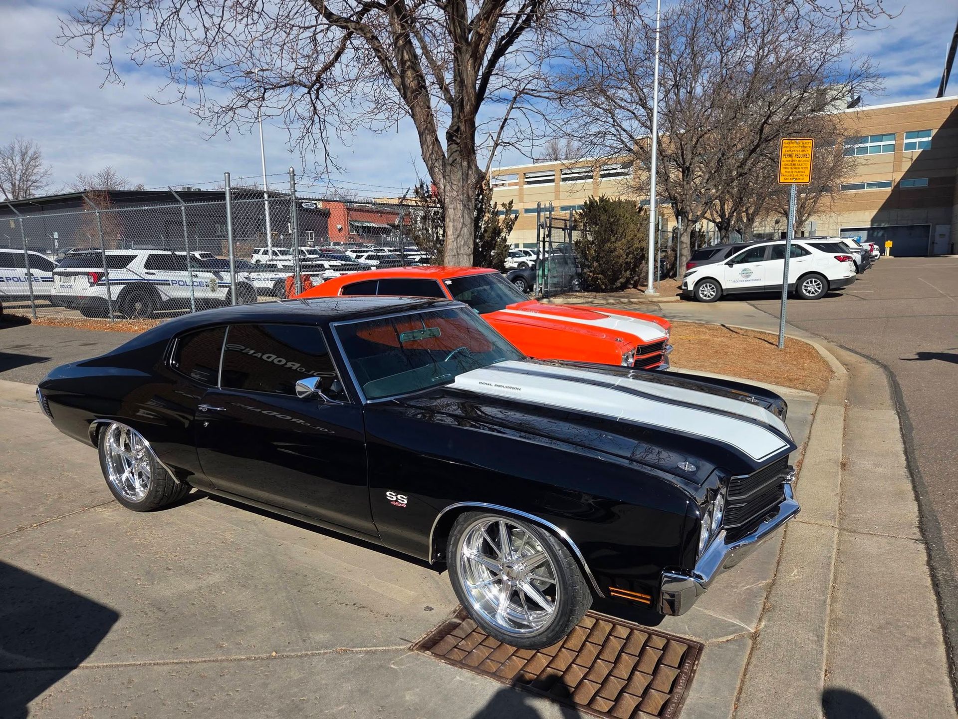 Black classic muscle car with white racing stripes parked outdoors near a red car and building