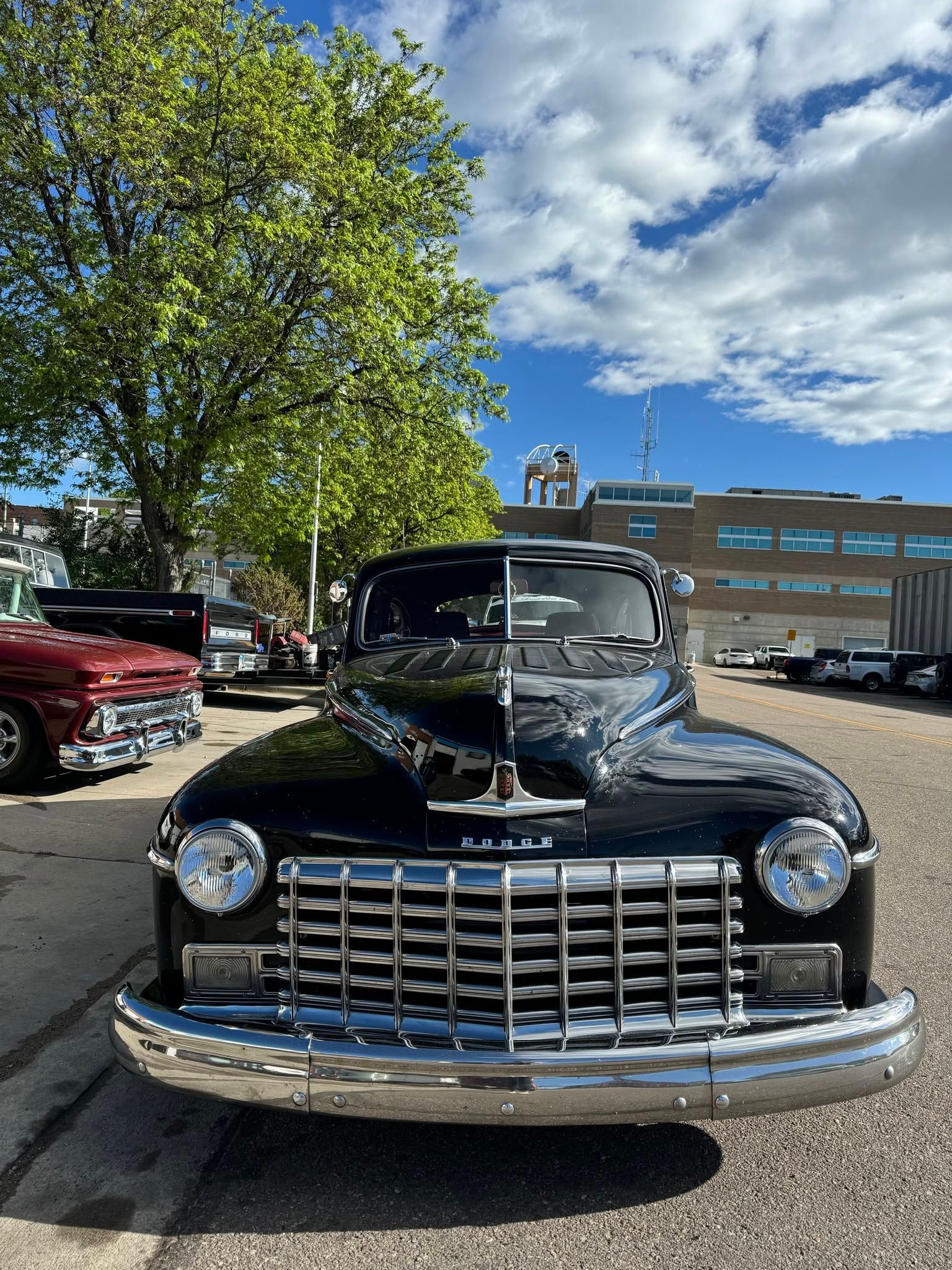 Front view of a black vintage car parked outdoors under a blue sky, with a red car and building behind it