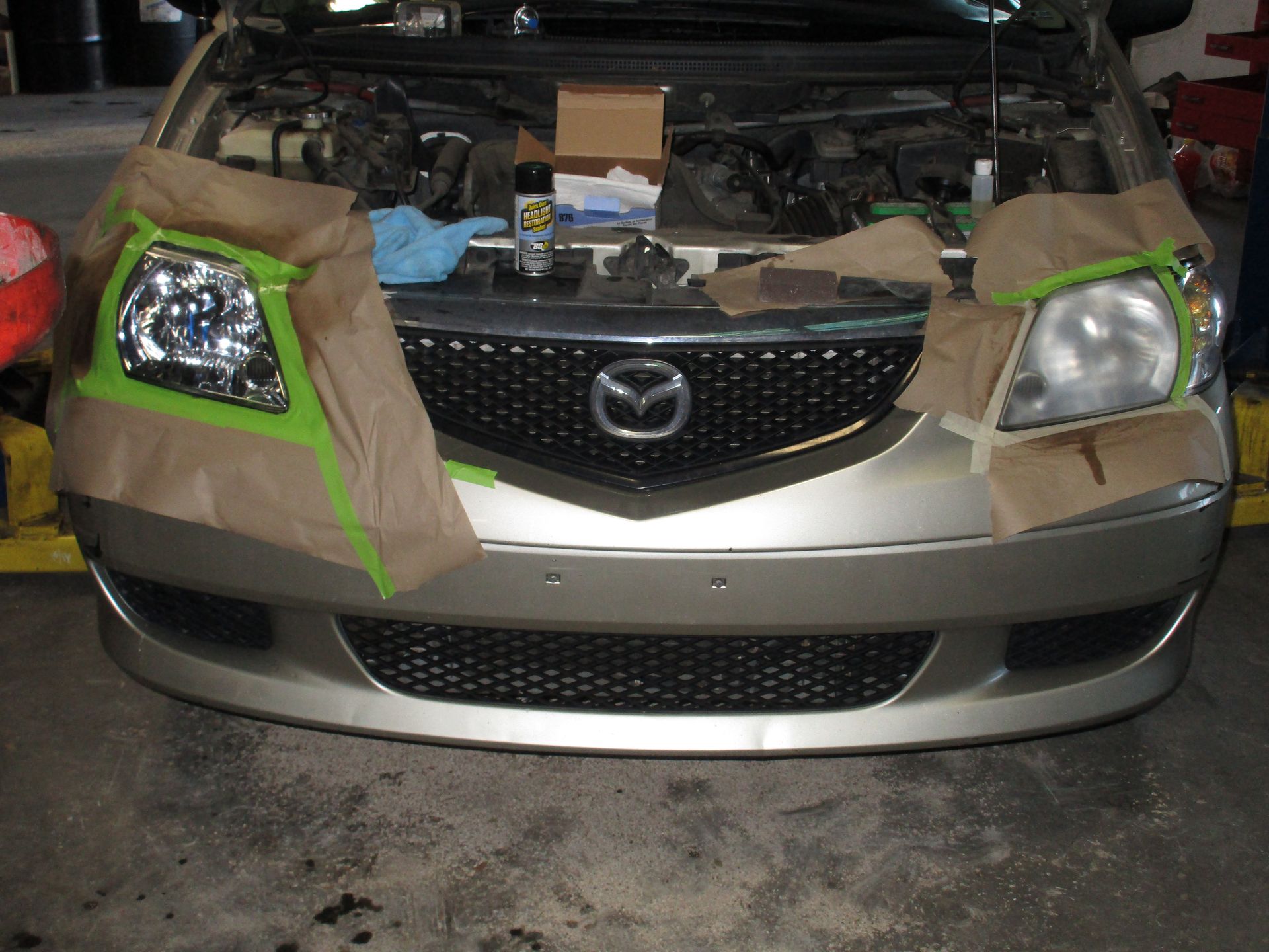 Front view of a gold Mazda with taped-off headlights in a garage, hood open and parts visible