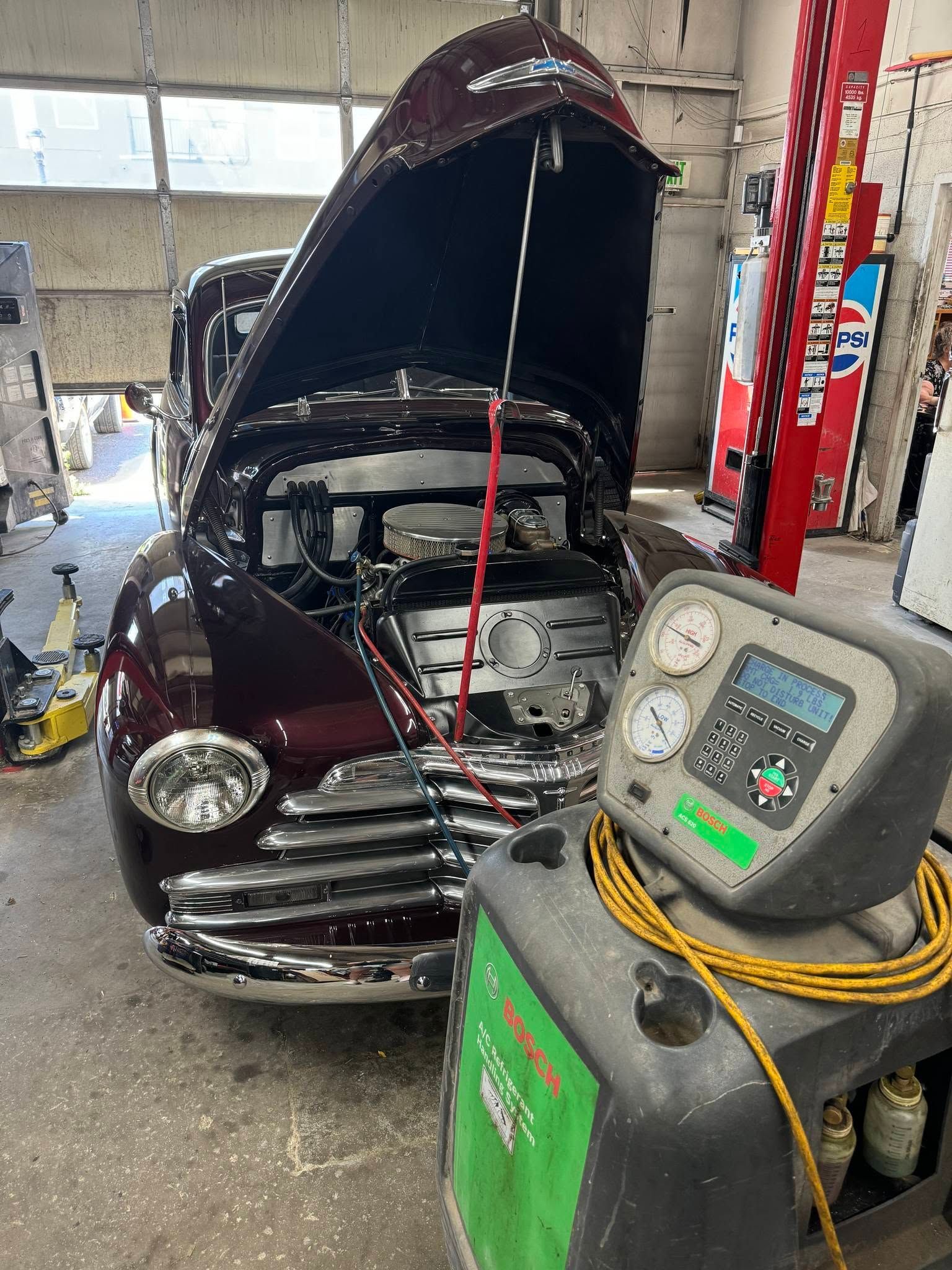 Classic car with hood open in an auto shop, showing engine work beside a tire machine.