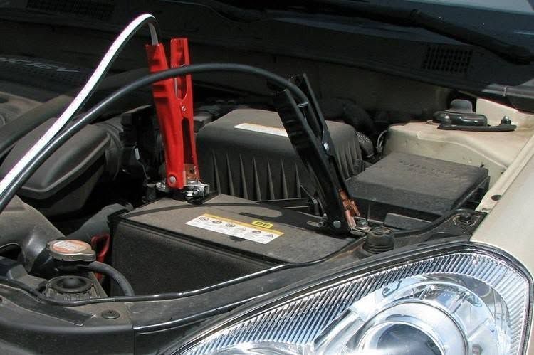 Car battery being jump-started in a car engine bay with red and black jumper cables.