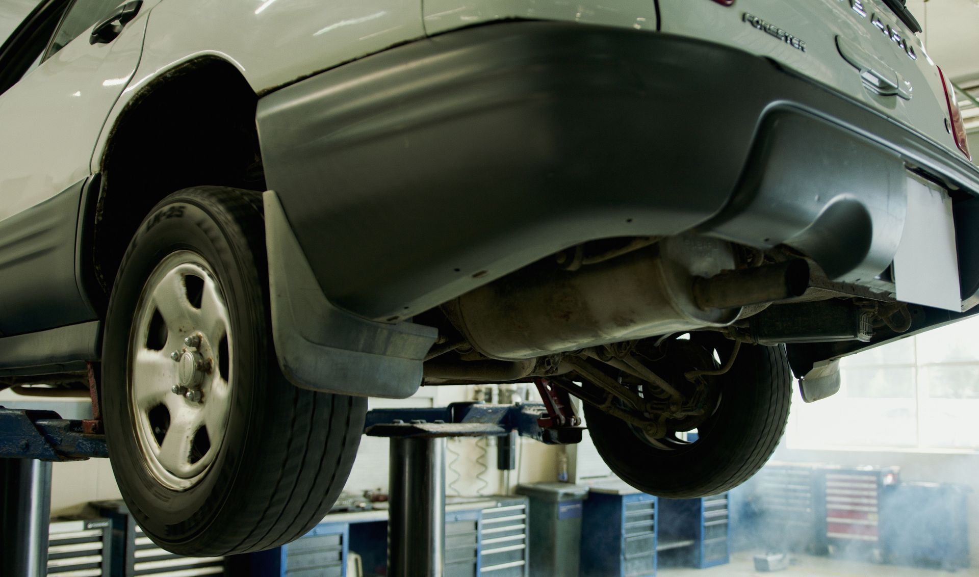 Underside of a gray SUV on a lift in an auto repair shop, with the rear wheel visible