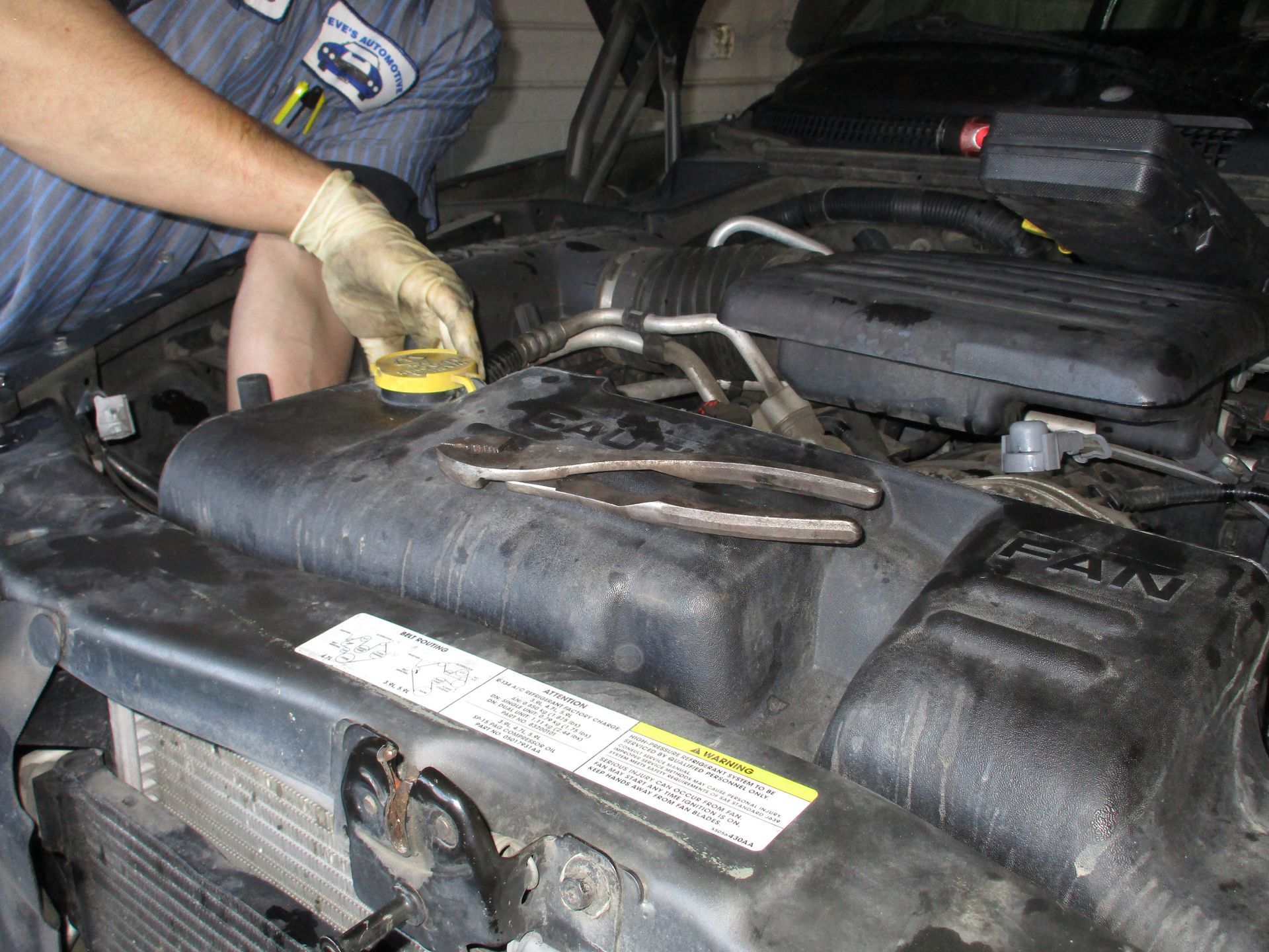 Mechanic working under a car hood, checking engine components with a yellow-gloved hand.