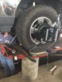 Mechanic under a lifted truck repairing a large off-road tire with a wheel alignment machine