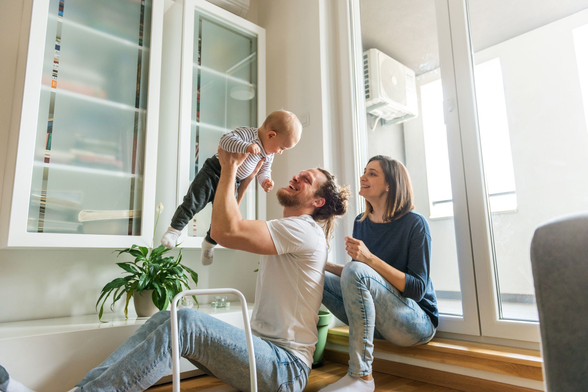 A couple sits on a couch and rug with their dog in a cozy, bright living room.