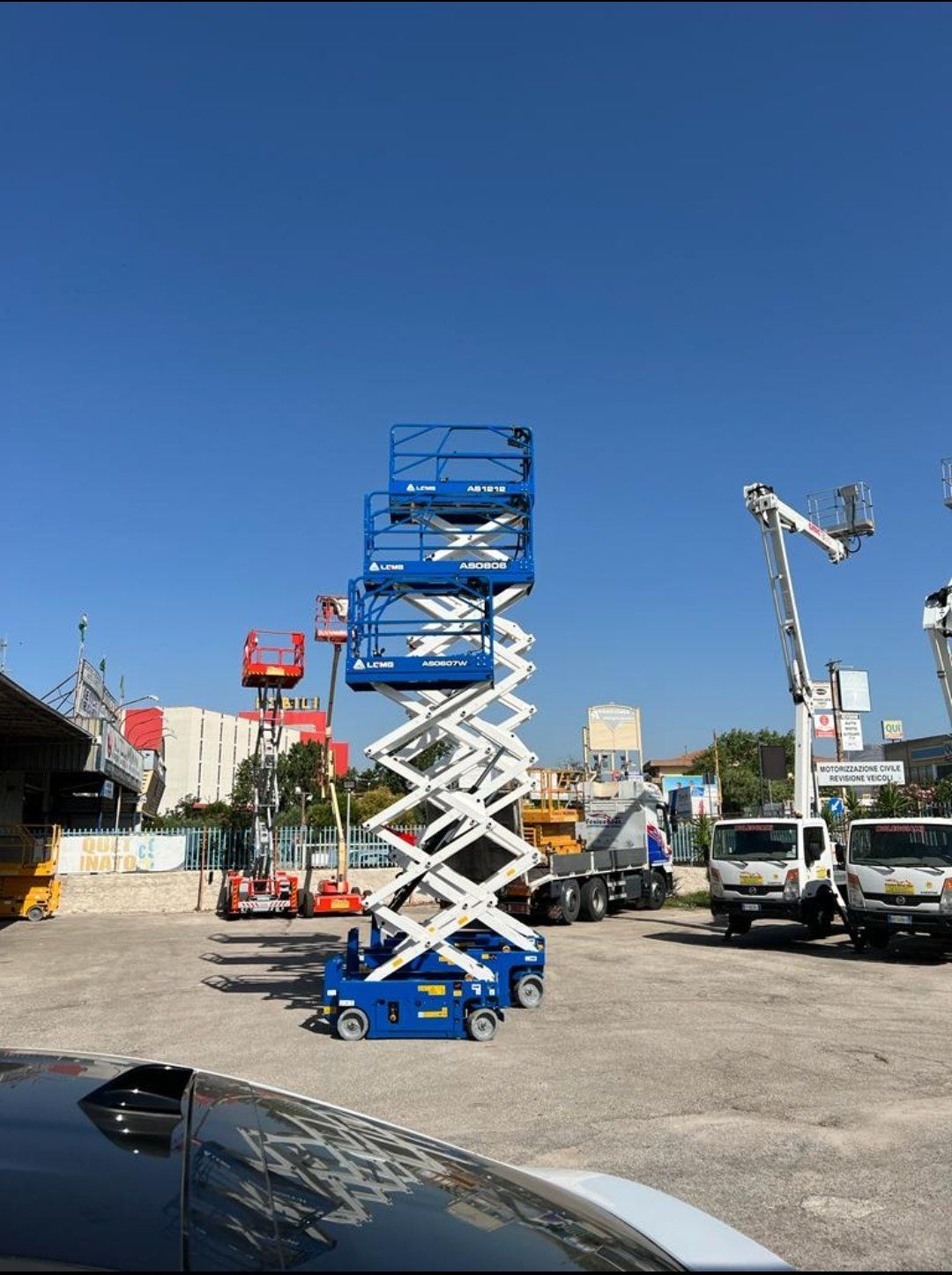 Piattaforma aerea a forbice blu in un deposito di attrezzature sotto un cielo terso. Sono visibili altri sollevatori e camion.