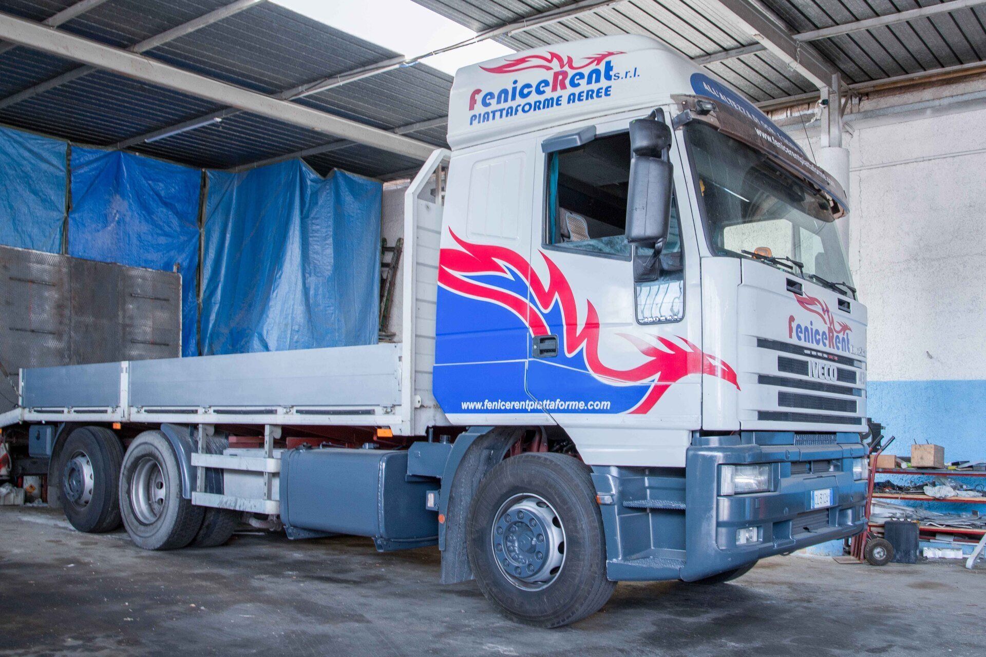Camion con pianale, cabina bianca con dettagli rossi e blu, all'interno di un magazzino. Logo 
