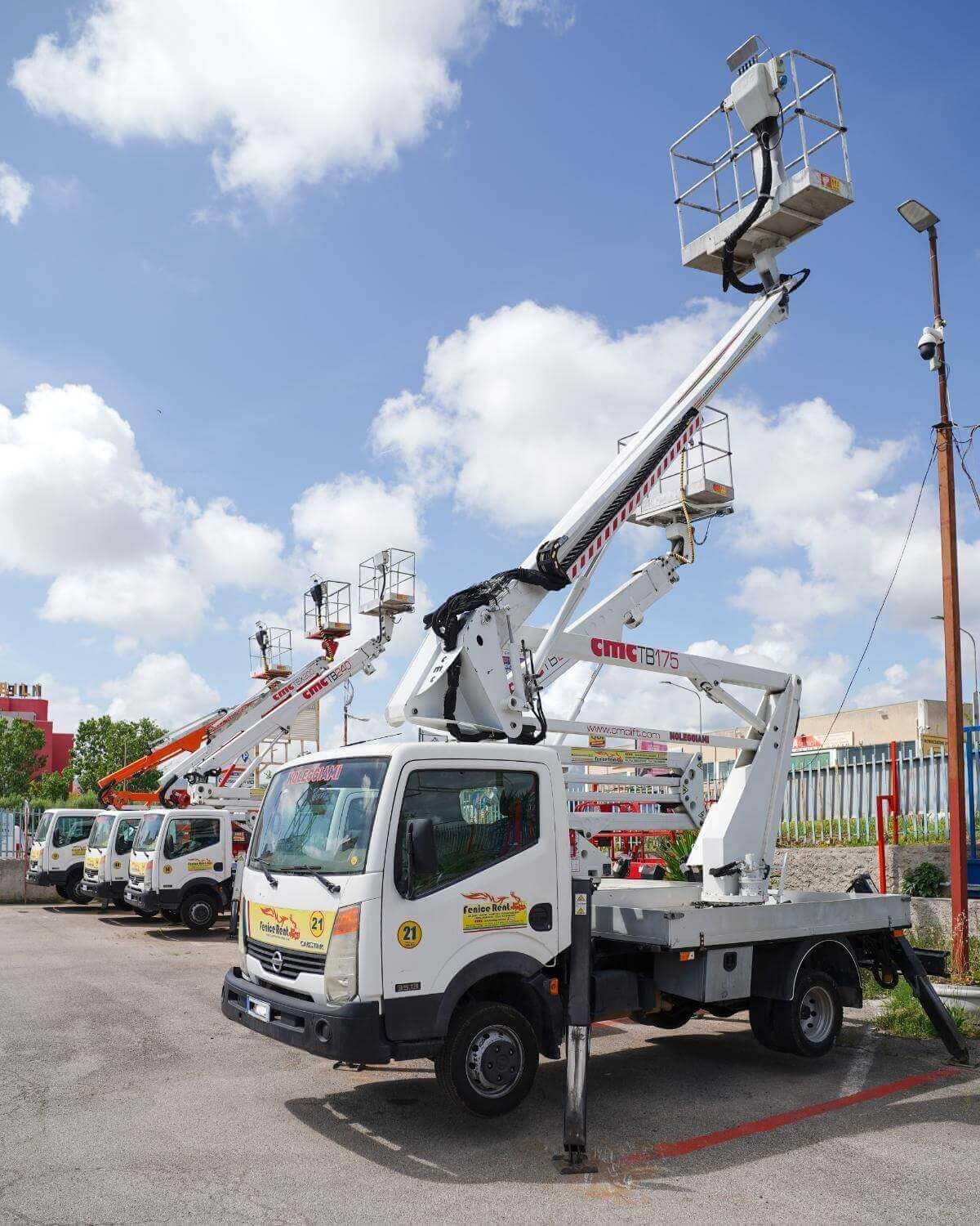 Un camion bianco con una piattaforma aerea sollevata in un parcheggio sotto un cielo nuvoloso. Altri camion simili si vedono sullo sfondo.