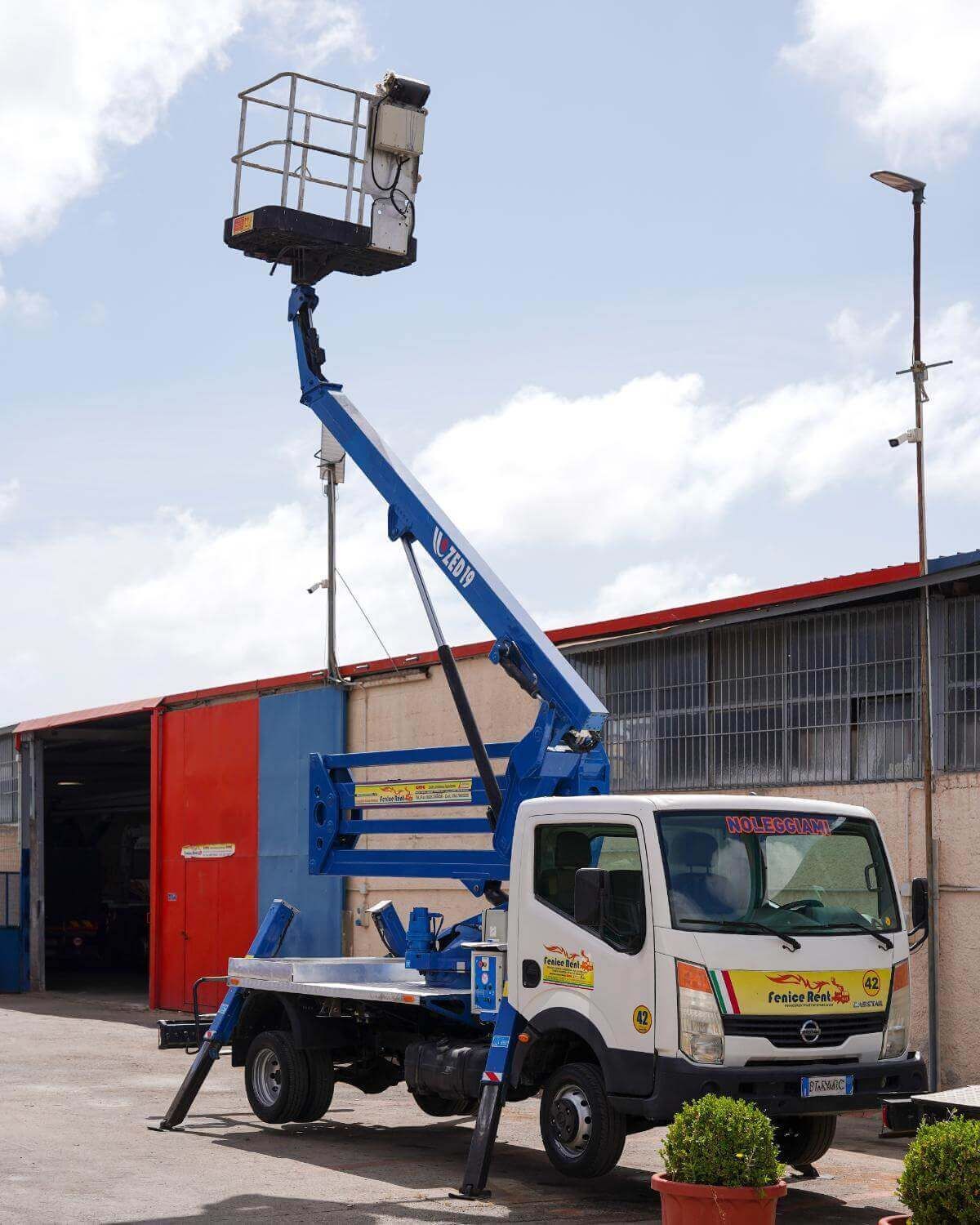 Piattaforma aerea montata su camion con braccio blu esteso, cestello in alto, gambe stabilizzatrici distese, parcheggiata vicino a un edificio, cielo nuvoloso.