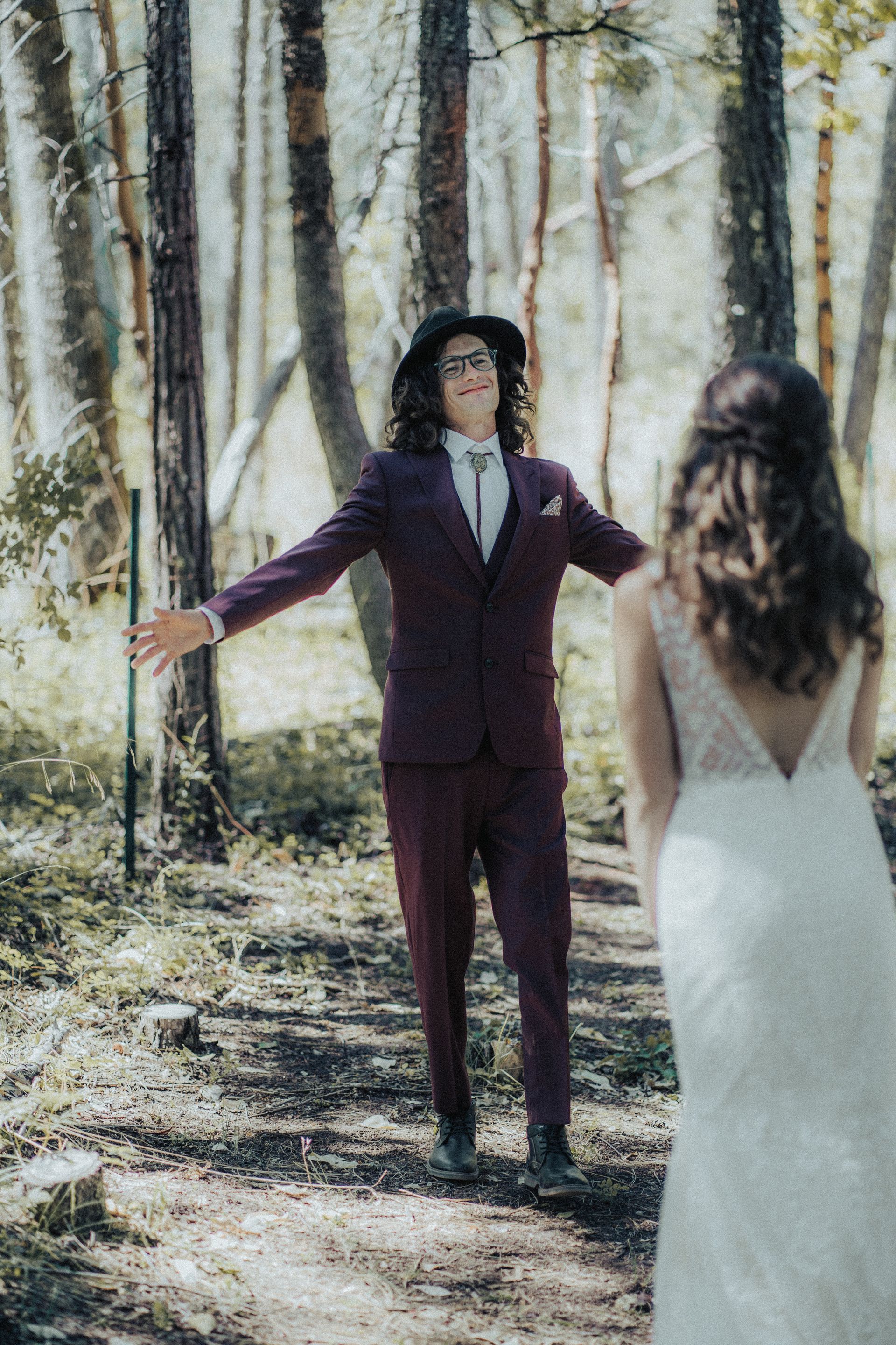 A bride and groom are standing in the woods . the groom is wearing a purple suit and a black hat.