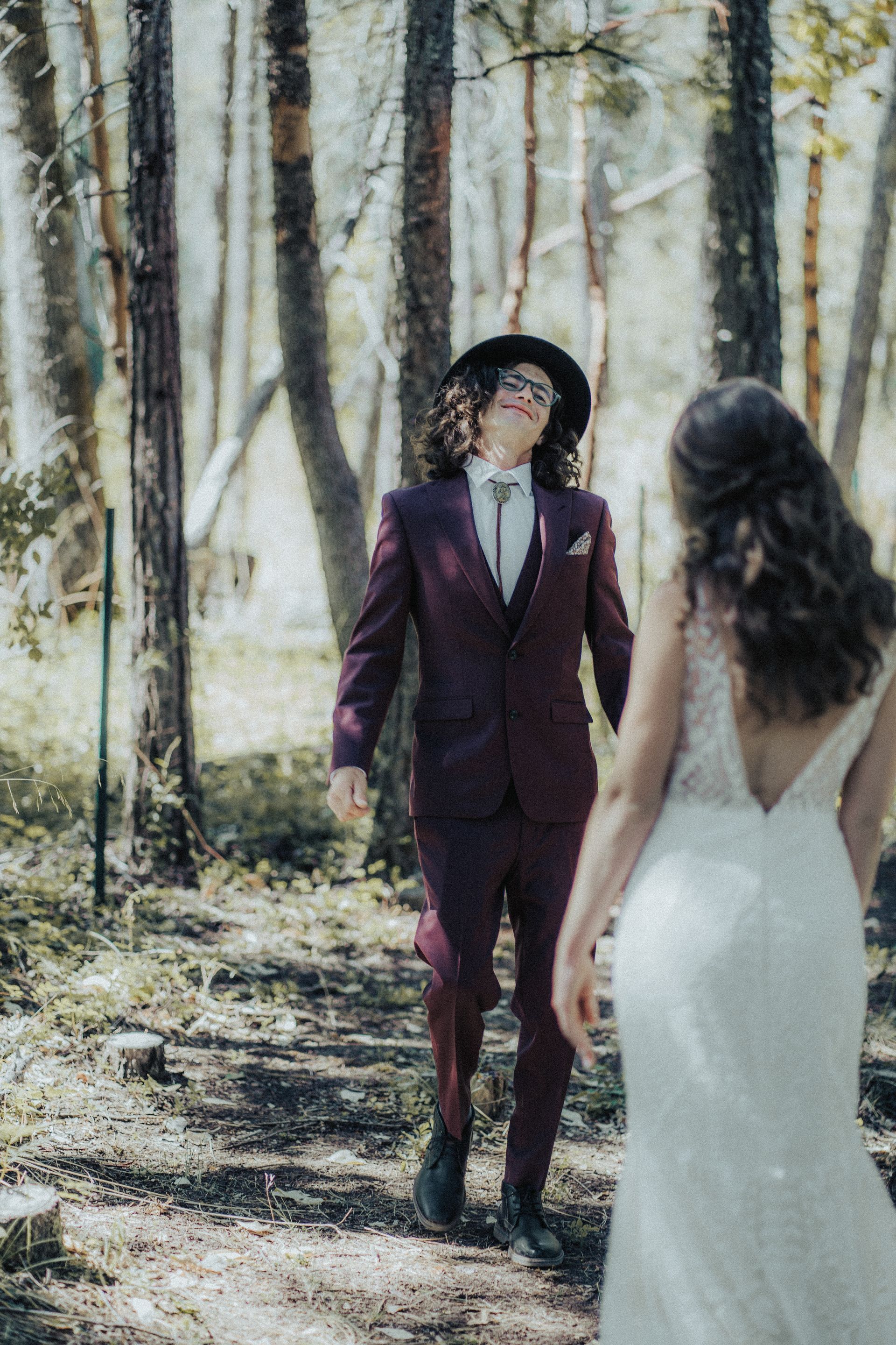 A bride and groom are walking through the woods holding hands.
