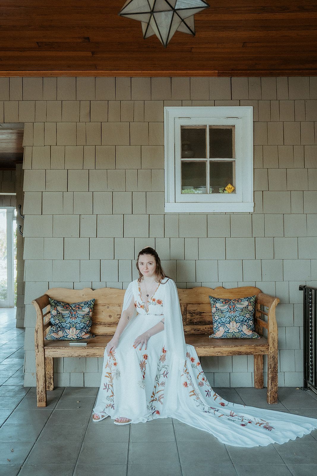 Woman in white dress with floral embroidery sits on a wooden bench, outdoors.