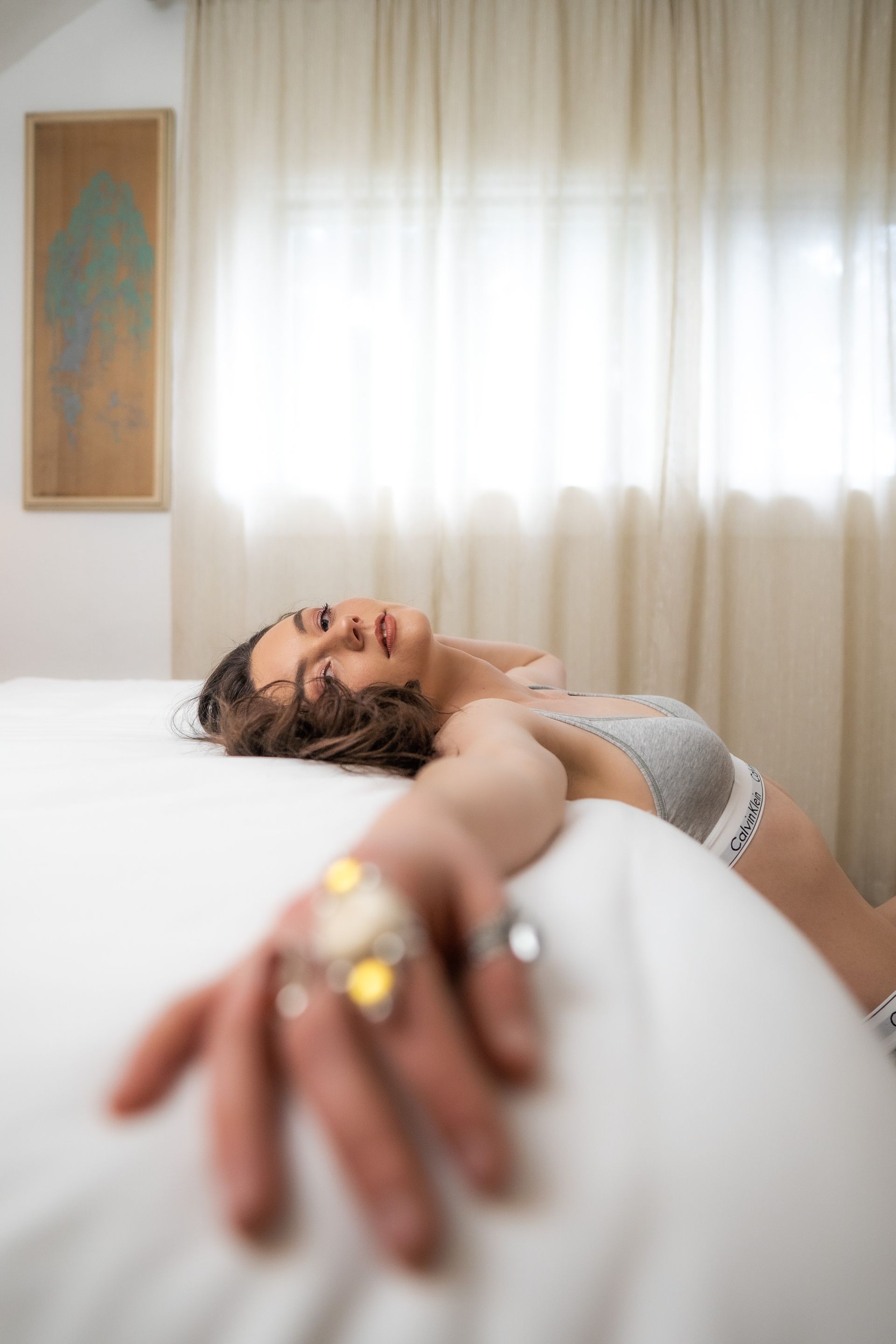 A woman with a grey bra on is leaning up against a bed for a boudoir photo shoot.