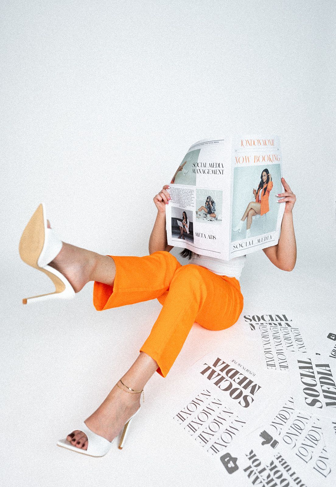 A woman is sitting on the floor holding a newspaper in front of her face.