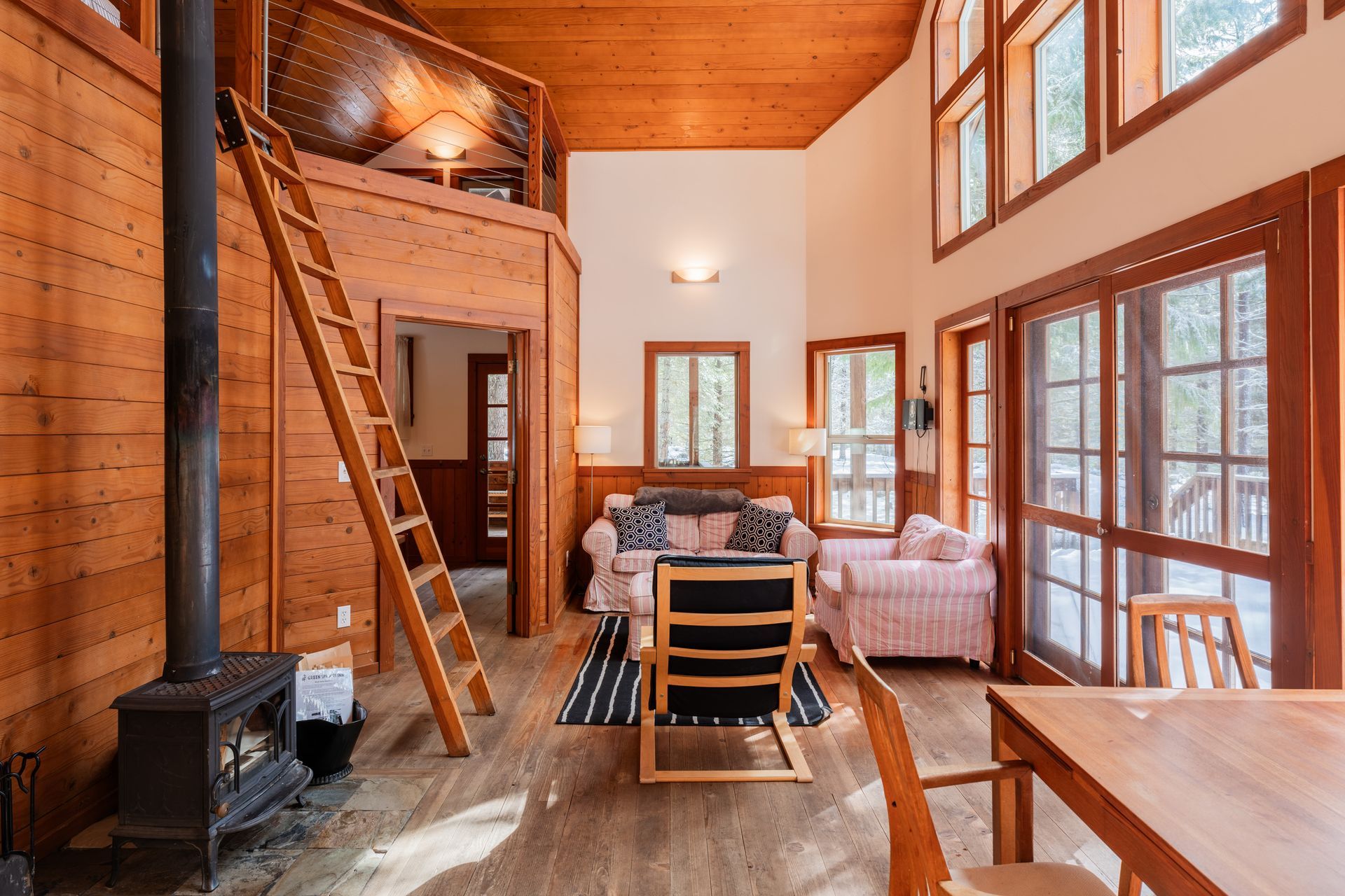 A living room in a log cabin with a wood stove , chairs , a table and a ladder.