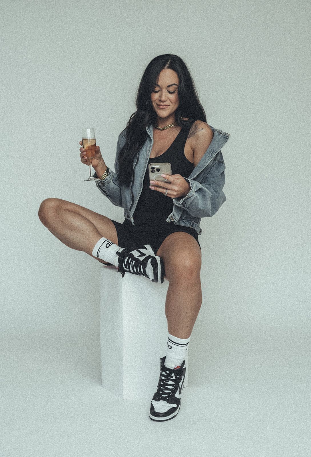 A woman is sitting on a cube holding a glass of wine and a cell phone.