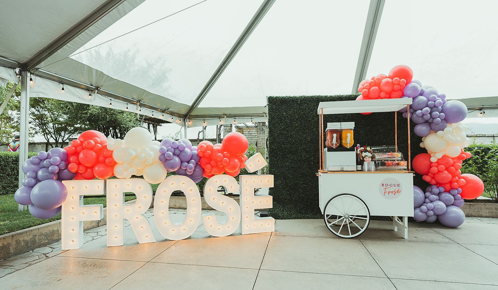 A sign that says `` frose '' is surrounded by balloons and a cart.