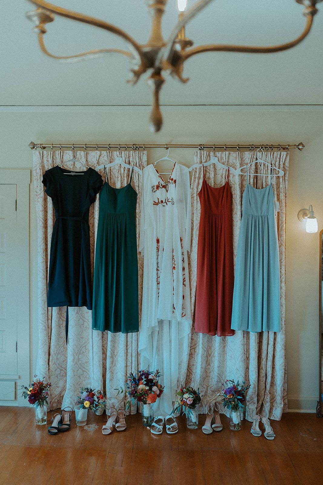 Five colorful bridesmaid dresses hanging on a curtained wall, below a chandelier.