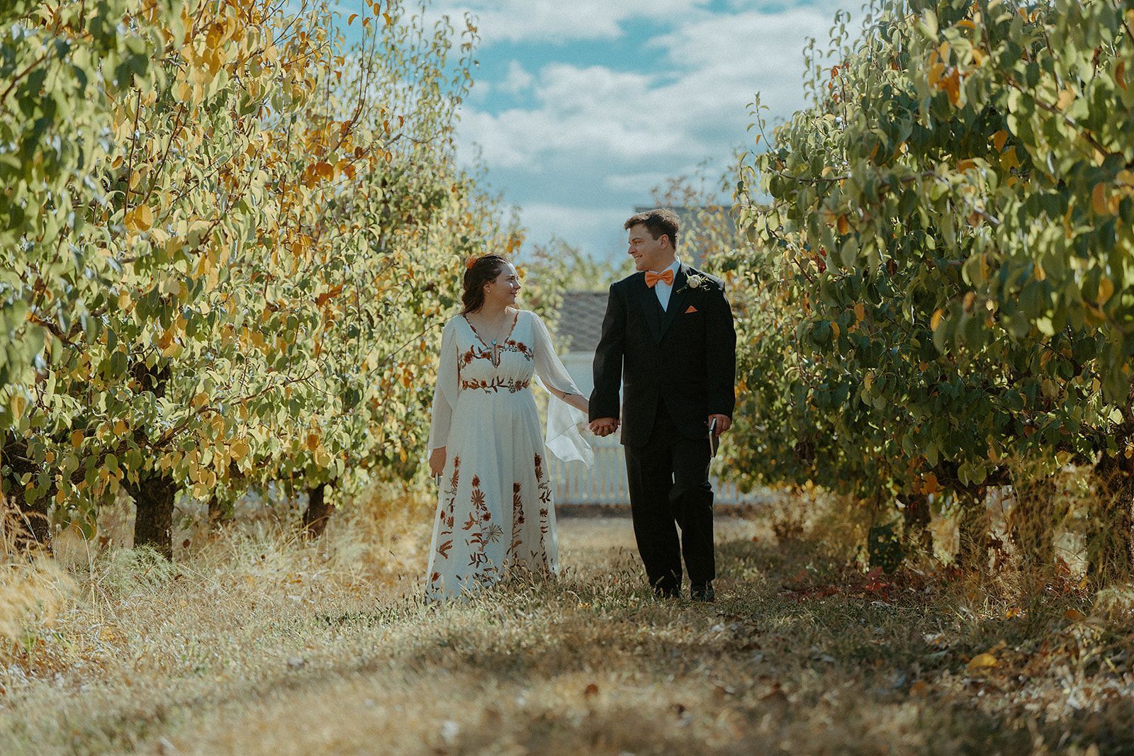 Bride and groom holding hands, walking down a path between trees, smiling, on a sunny day.