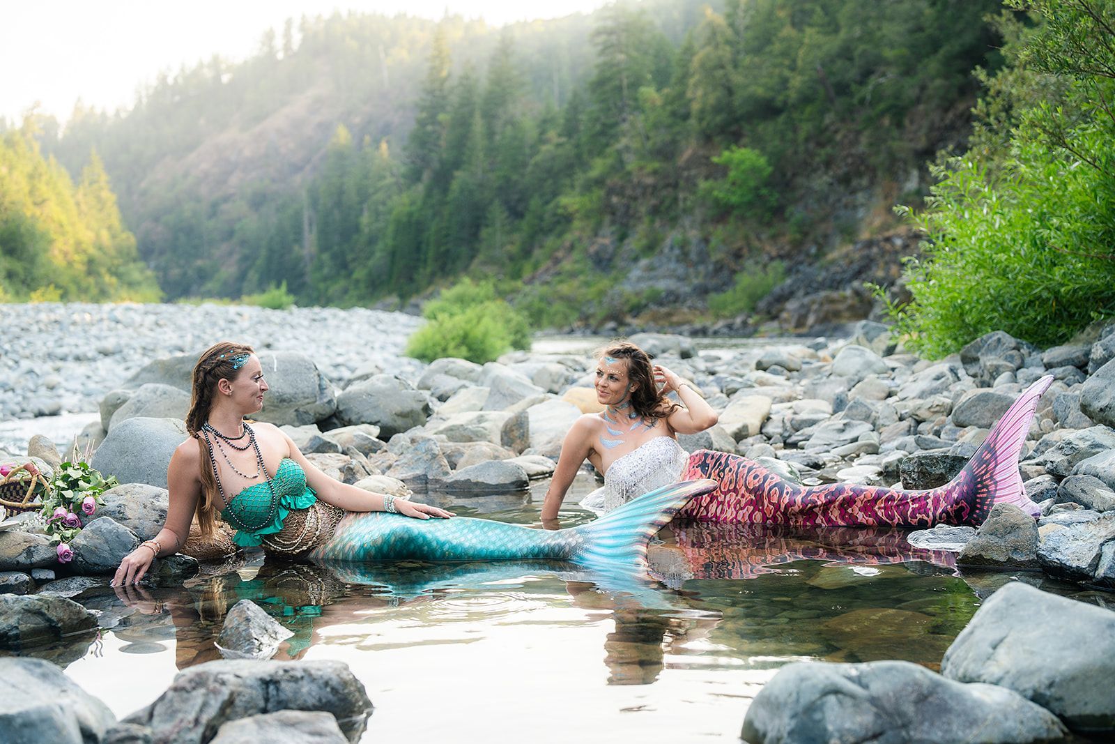 Two mermaids are sitting in a river surrounded by rocks.