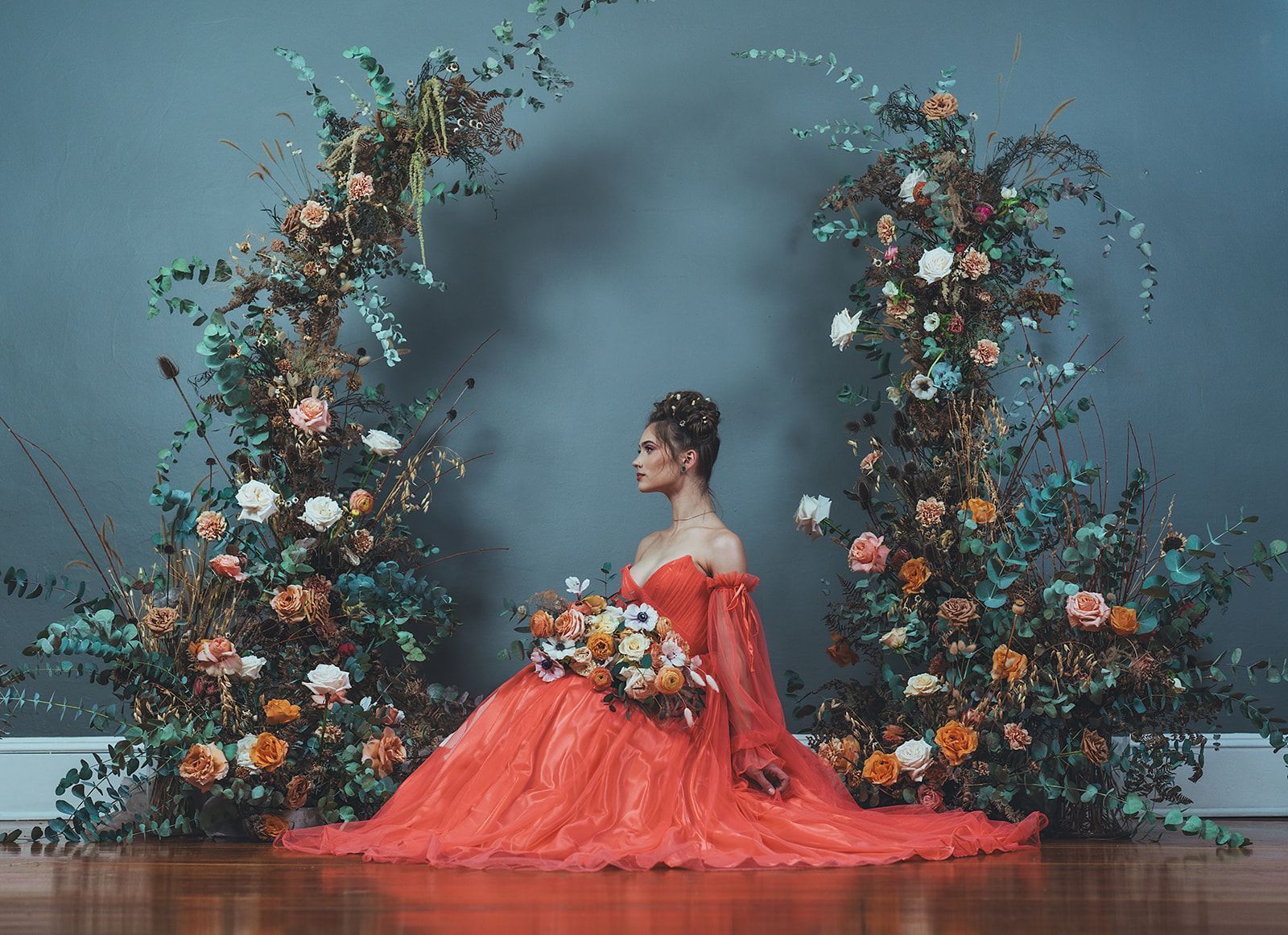 A woman in a red dress is sitting in front of a floral arch.