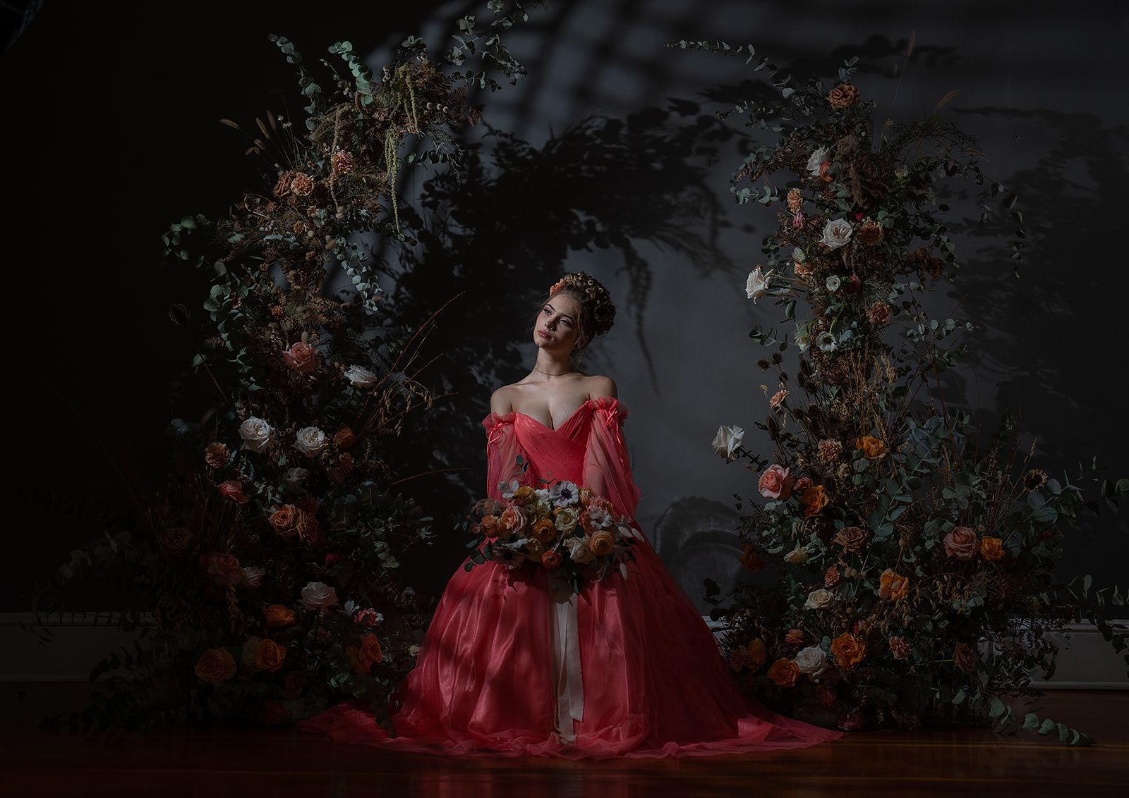 A woman in a red dress is standing in front of a wall of flowers.