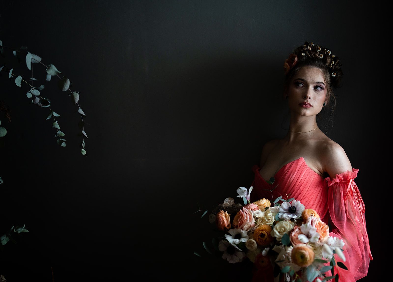 Woman in coral gown holding flowers, looking up, dark background.