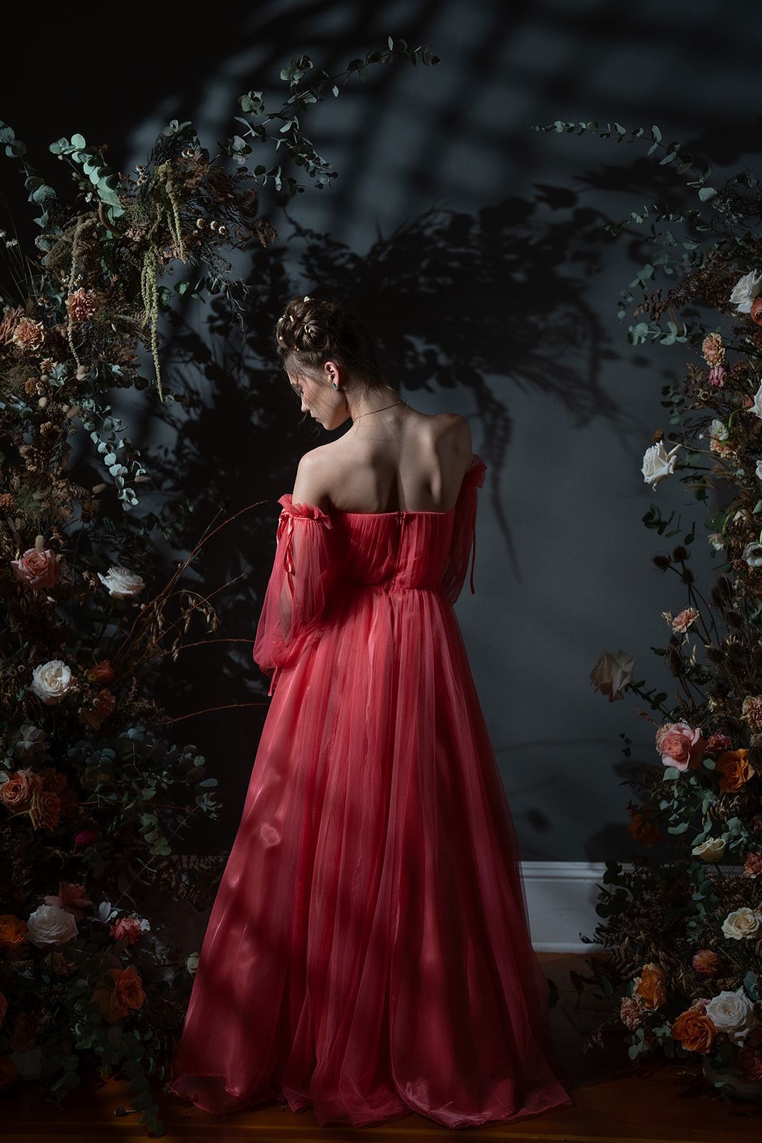 A woman in a red dress is standing in front of a wall of flowers.