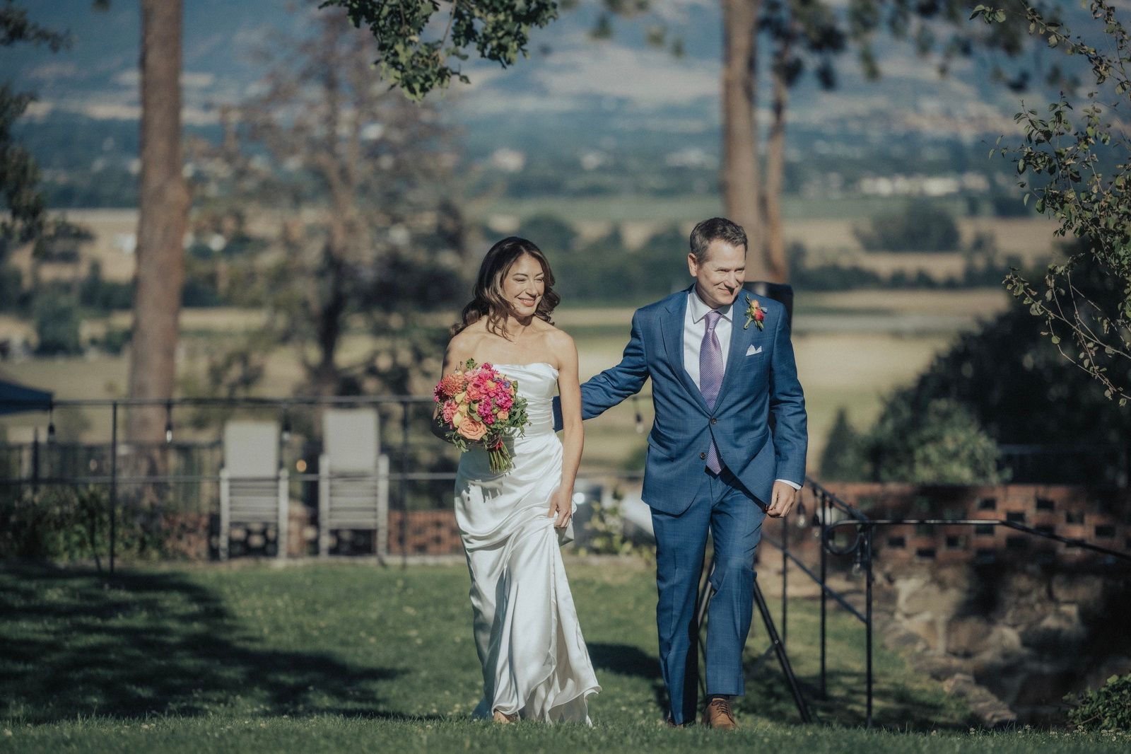 Bride and groom walk together after ceremony, smiling, outdoors with scenic background.