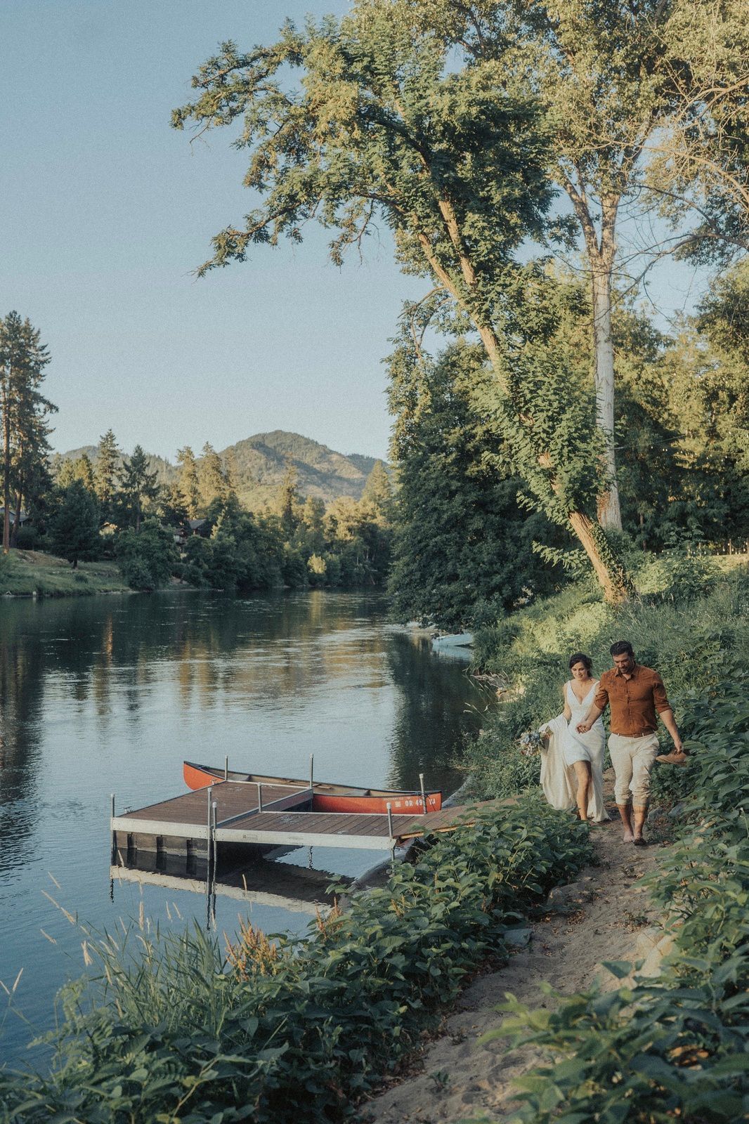 Couple walks along a river path, holding a blanket. Red canoe docked at a pier. Green trees and mountain.