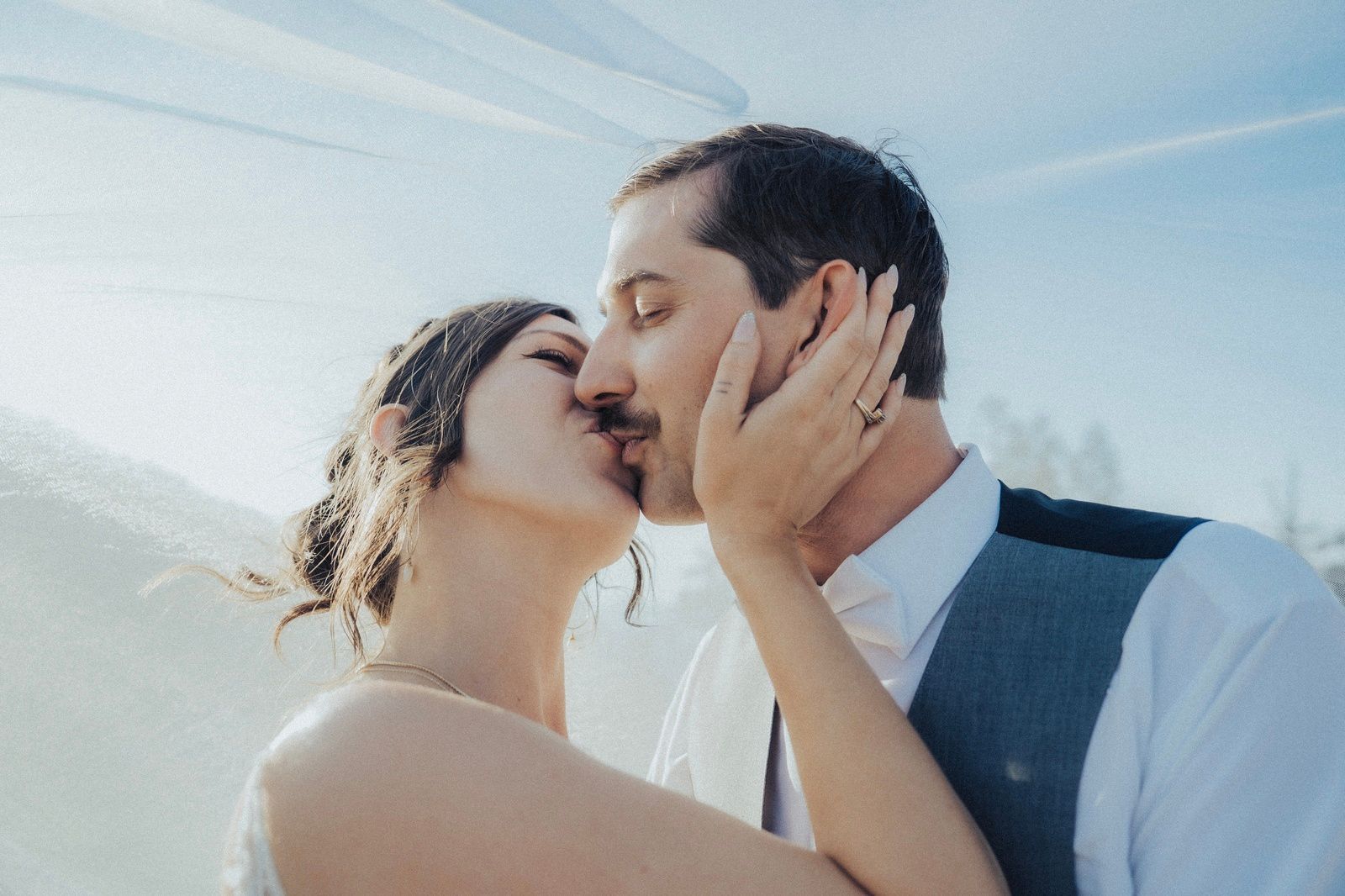 Couple kissing, woman's hand on man's face. Wedding, outdoors, veil, bright sky.