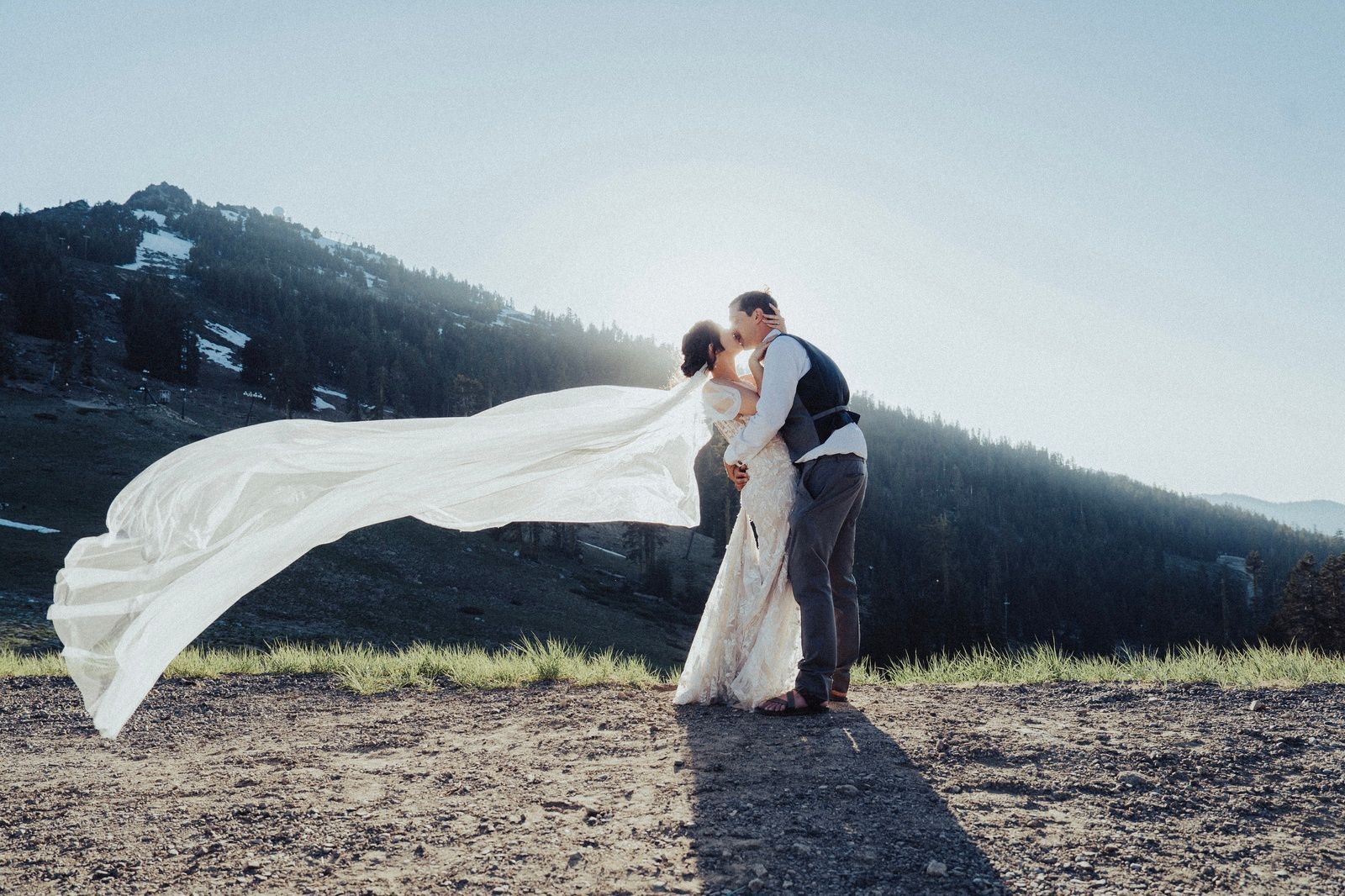 Couple kissing in sunlight with a long veil flowing in the wind, mountain backdrop.
