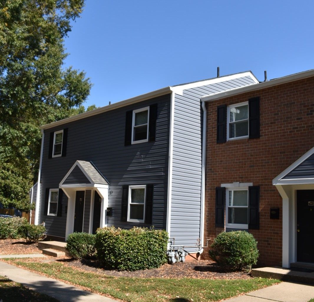 Two-story apartment buildings with gray siding and red brick, front stoops, and shrubs under a clear blue sky