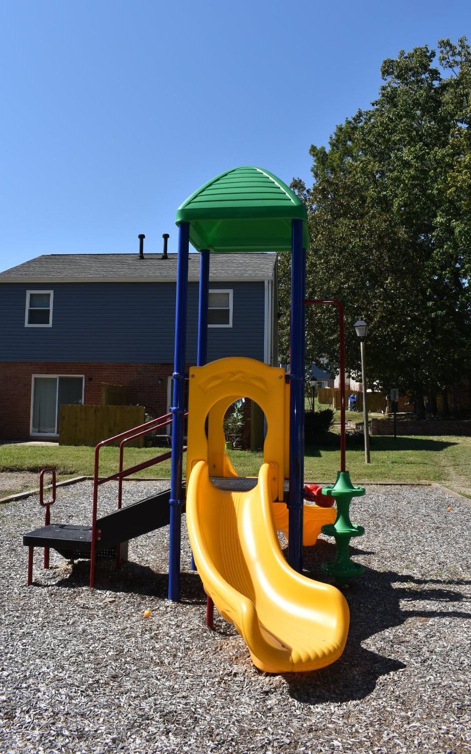 Yellow playground slide with green roof and blue support posts in a gravel play area near a house.