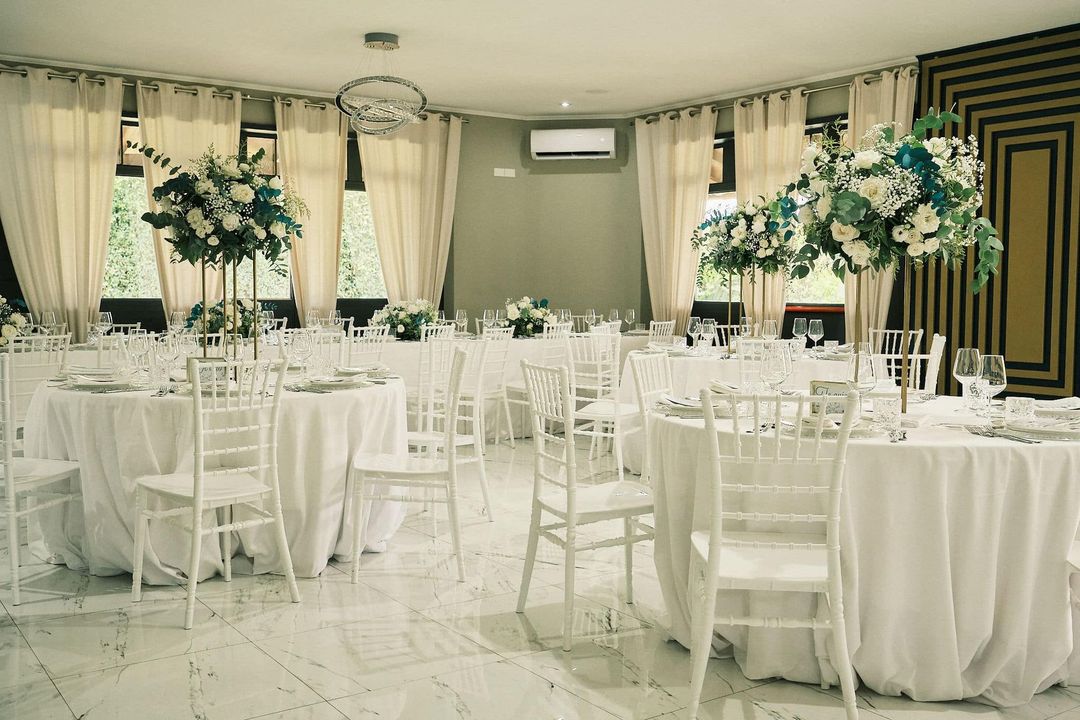 Elegante sala per banchetti allestita con sedie e tavoli bianchi, decorata con centrotavola floreali verdi e bianchi.