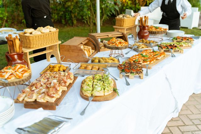 Tavolo da buffet all'aperto con assortimento di pasticcini, pane e antipasti su una tovaglia bianca.