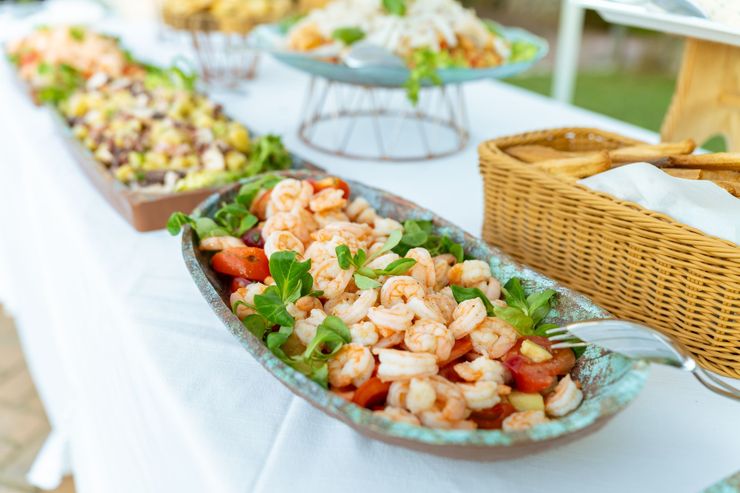 Tavolo da buffet con insalata di mare in una ciotola di vetro, altre pietanze e un cestino del pane su una tovaglia bianca.