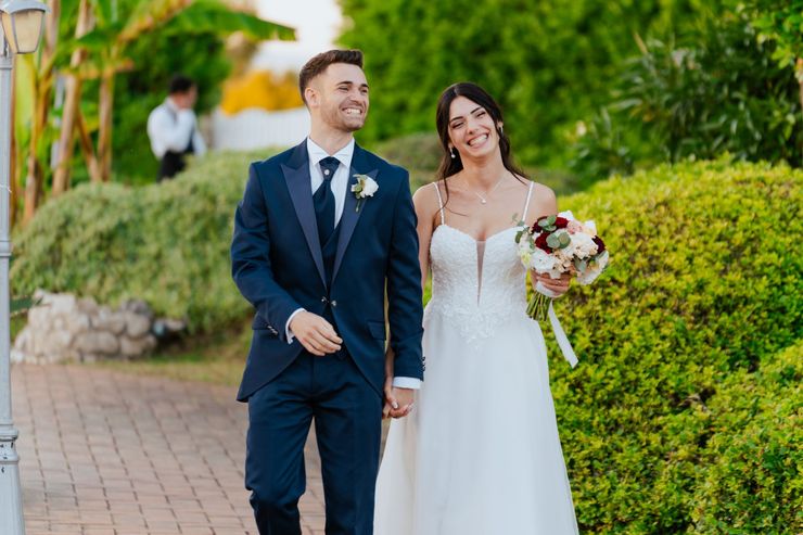 Gli sposi camminano mano nella mano in un giardino, sorridenti, con i bouquet e gli abiti da cerimonia.