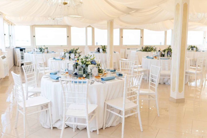Elegante sala ricevimenti bianca con tavoli rotondi, sedie bianche e centrotavola floreali sotto drappeggi di tessuto.