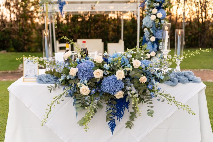 Tavolo da ricevimento bianco con composizione floreale blu e bianca e candele all'aperto