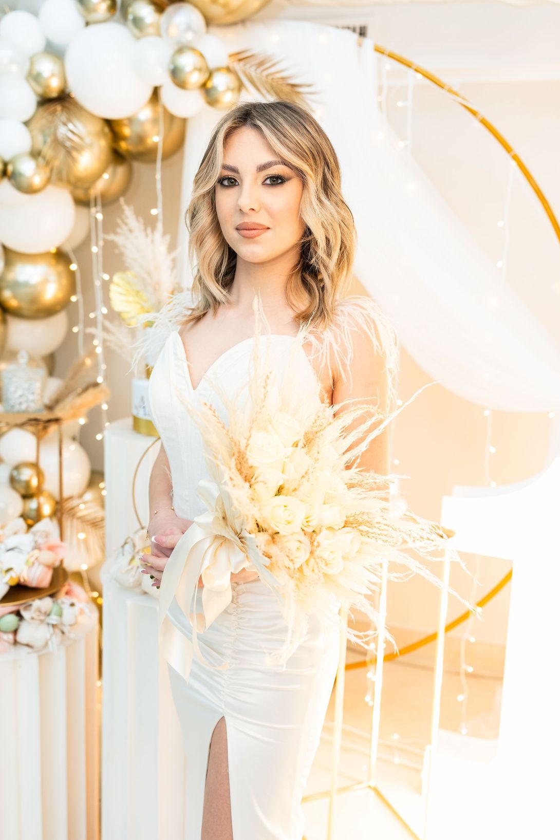 Donna in abito bianco che tiene in mano un bouquet dorato accanto a uno sfondo di palloncini dorati e bianchi.