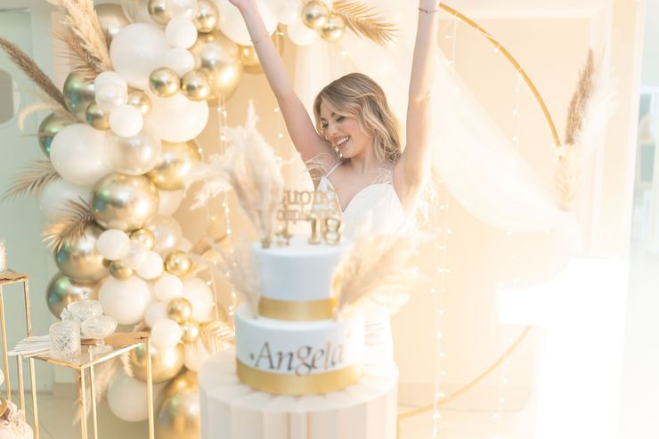 Donna accanto a una torta di compleanno bianca e oro con ghirlanda di palloncini e decorazioni floreali.